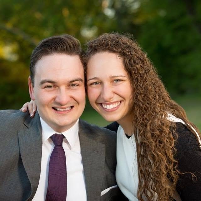 A smiling man in a gray suit and a woman with long curly hair in a black and white top, standing close together outdoors with greenery in the background.