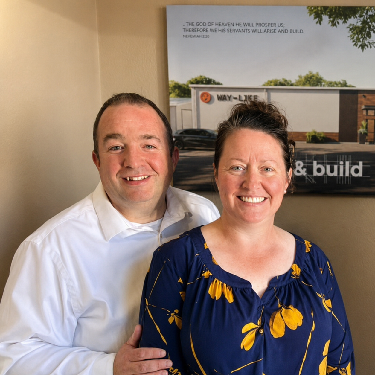 A smiling man in a white shirt standing next to a smiling woman in a blue and yellow floral blouse, with a framed picture and wall in the background.