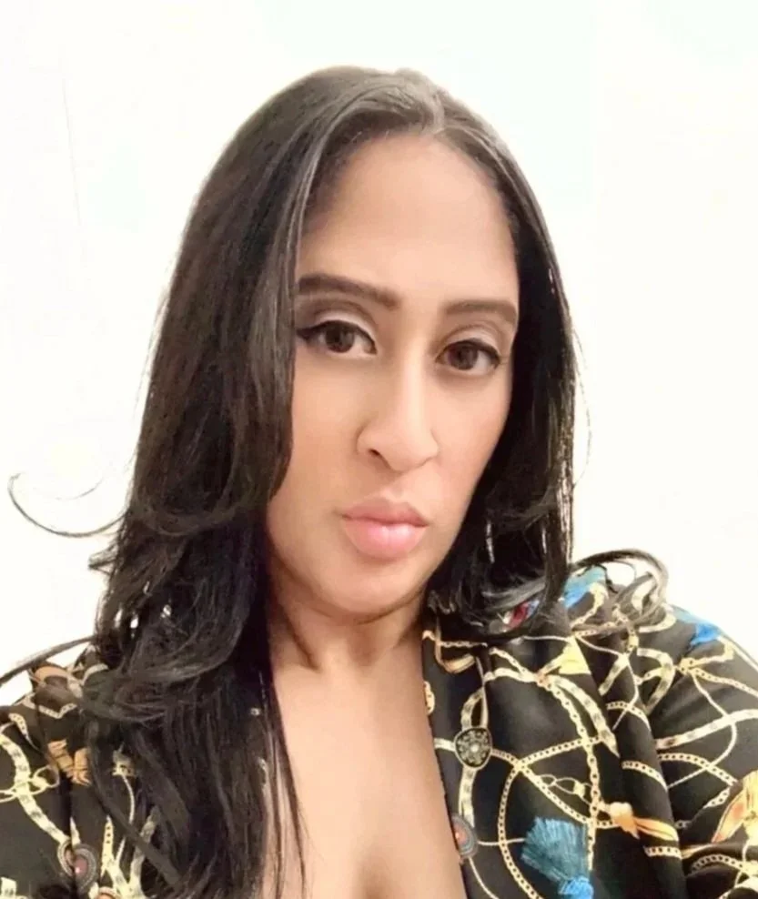 Portrait of a woman with long dark hair, wearing a black shirt with a chain and rope pattern, against a plain white background.