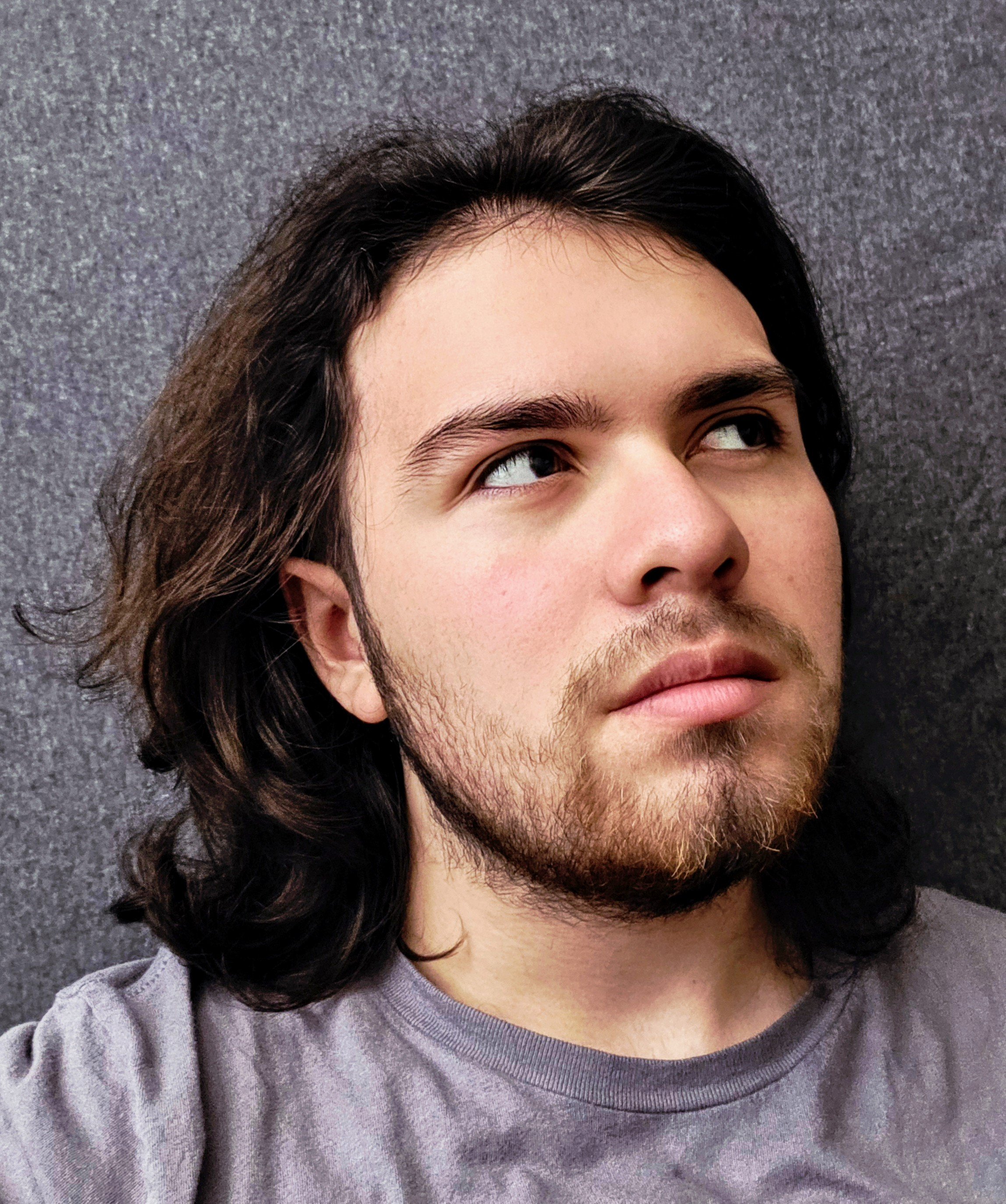Close-up of a person's face with light skin, dark brown hair, and a beard, looking upward, wearing a gray T-shirt, against a textured gray background.