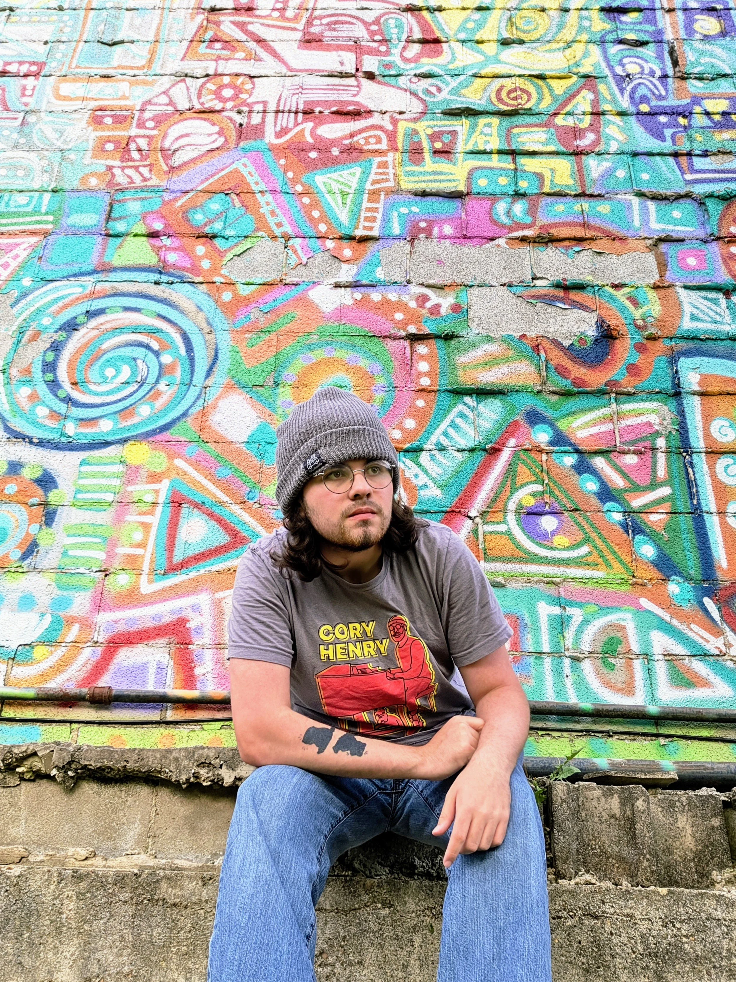 A young man with long brown hair, glasses, and a gray beanie, sitting on concrete steps in front of a colorful graffiti mural on a brick wall. The mural features abstract patterns and bright colors.