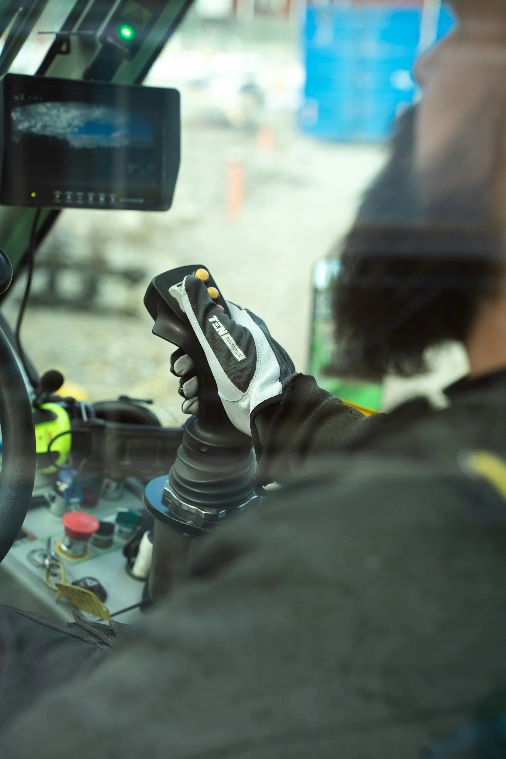 View inside a vehicle cab, showing a person operating machinery with a glove on their hand, control panels, and equipment, with a blurred outdoor scene through the window.