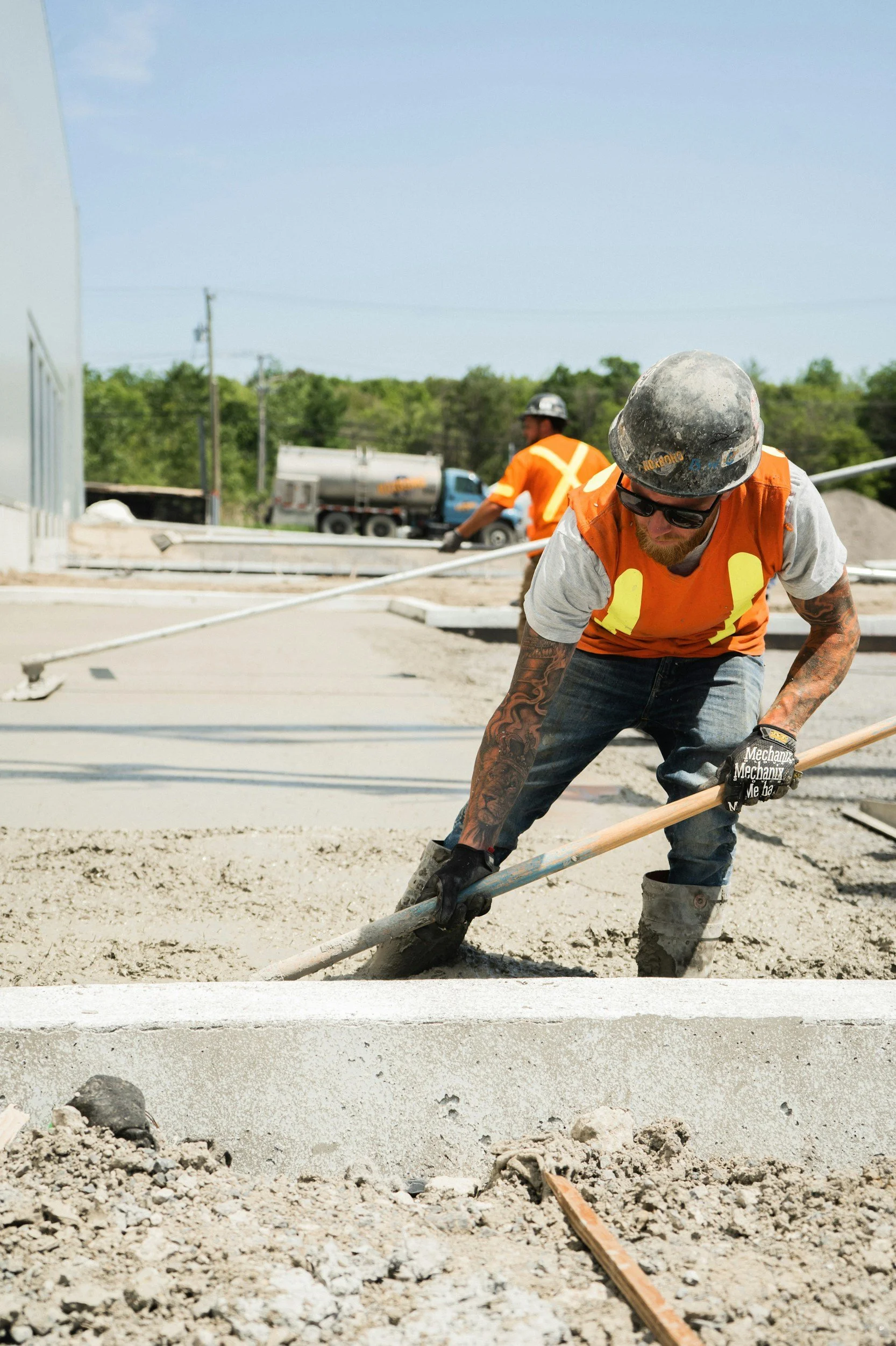Two construction workers in orange safety vests, hard hats, and sunglasses leveling concrete with a long-handled tool at an outdoor construction site.