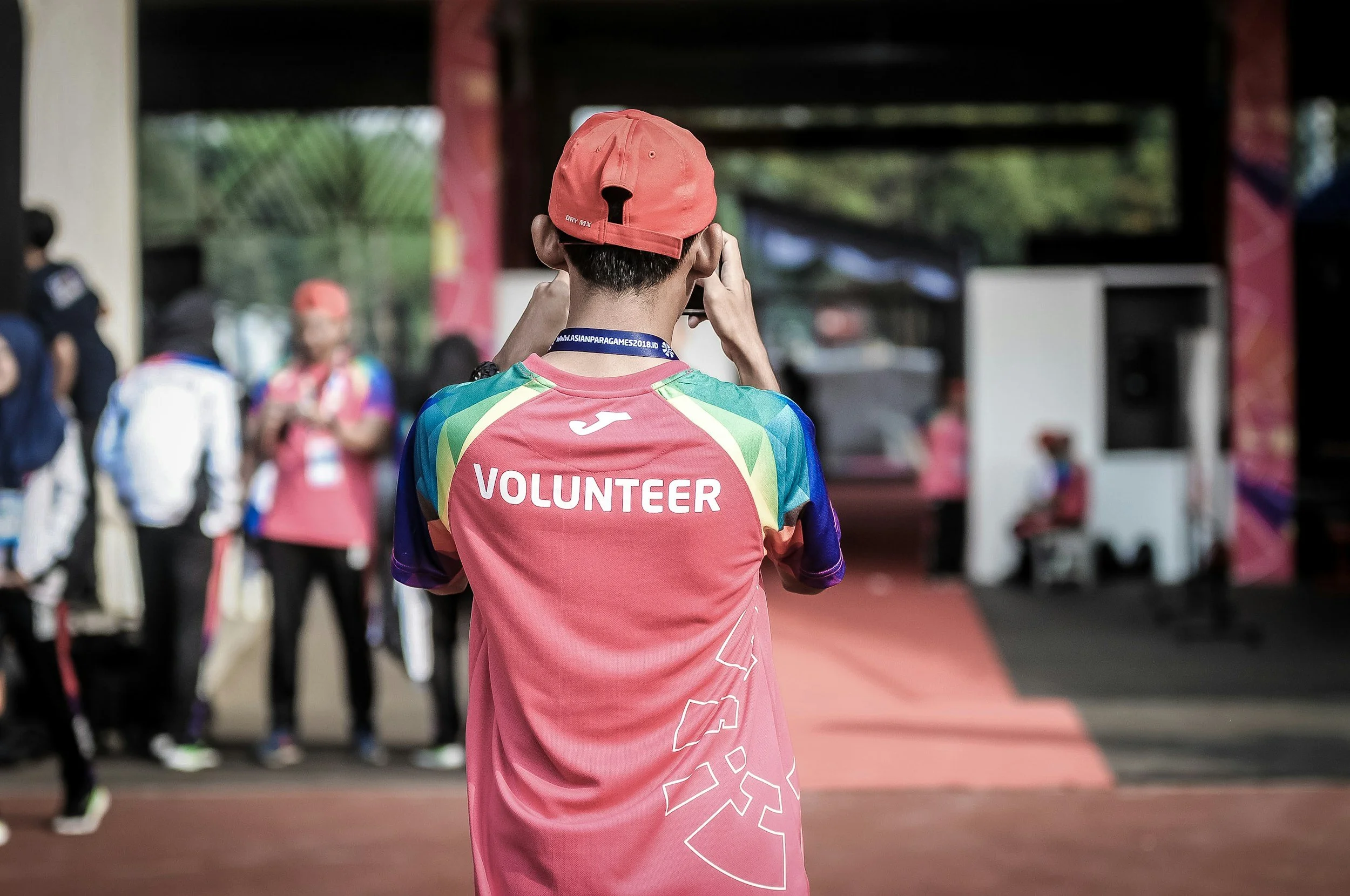 A person wearing a pink cap and colorful shirt with the word 'VOLUNTEER' on the back, standing in an indoor or partially covered area, with other people in the background at an event or gathering.