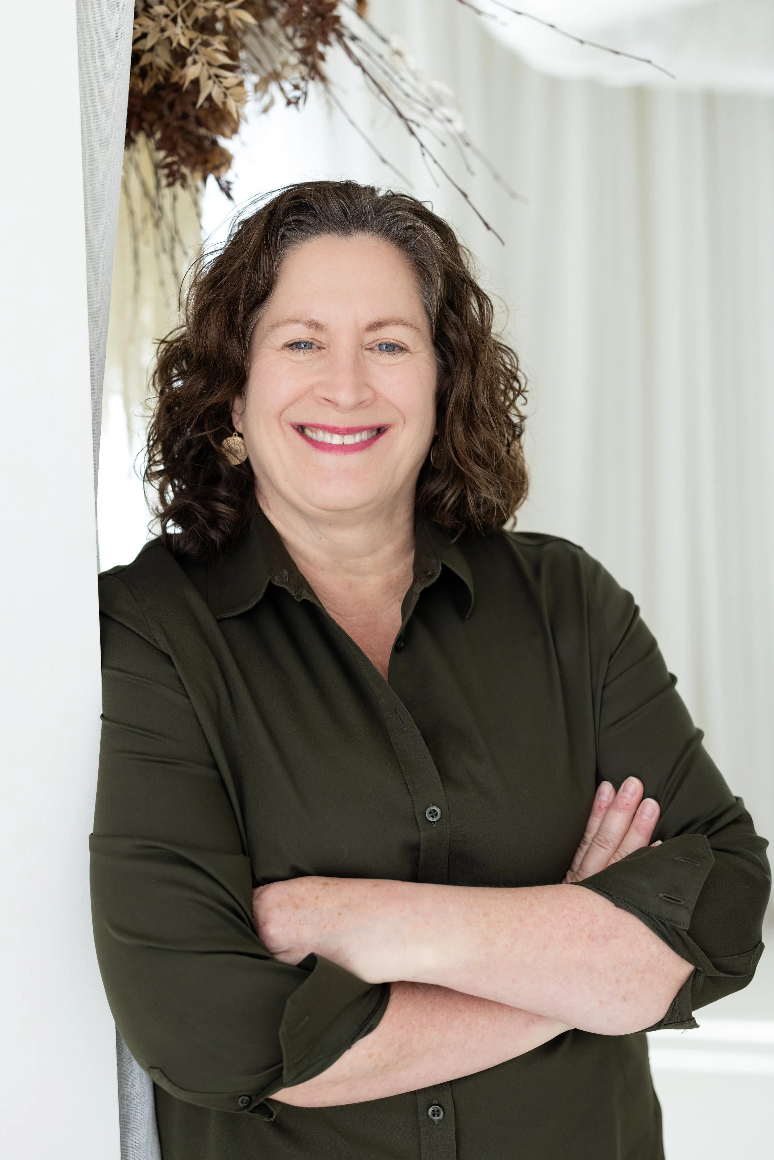 A woman with curly brown hair and wearing a dark green shirt, smiling with her arms crossed, standing in front of a light background with a dried floral arrangement in the upper left corner.