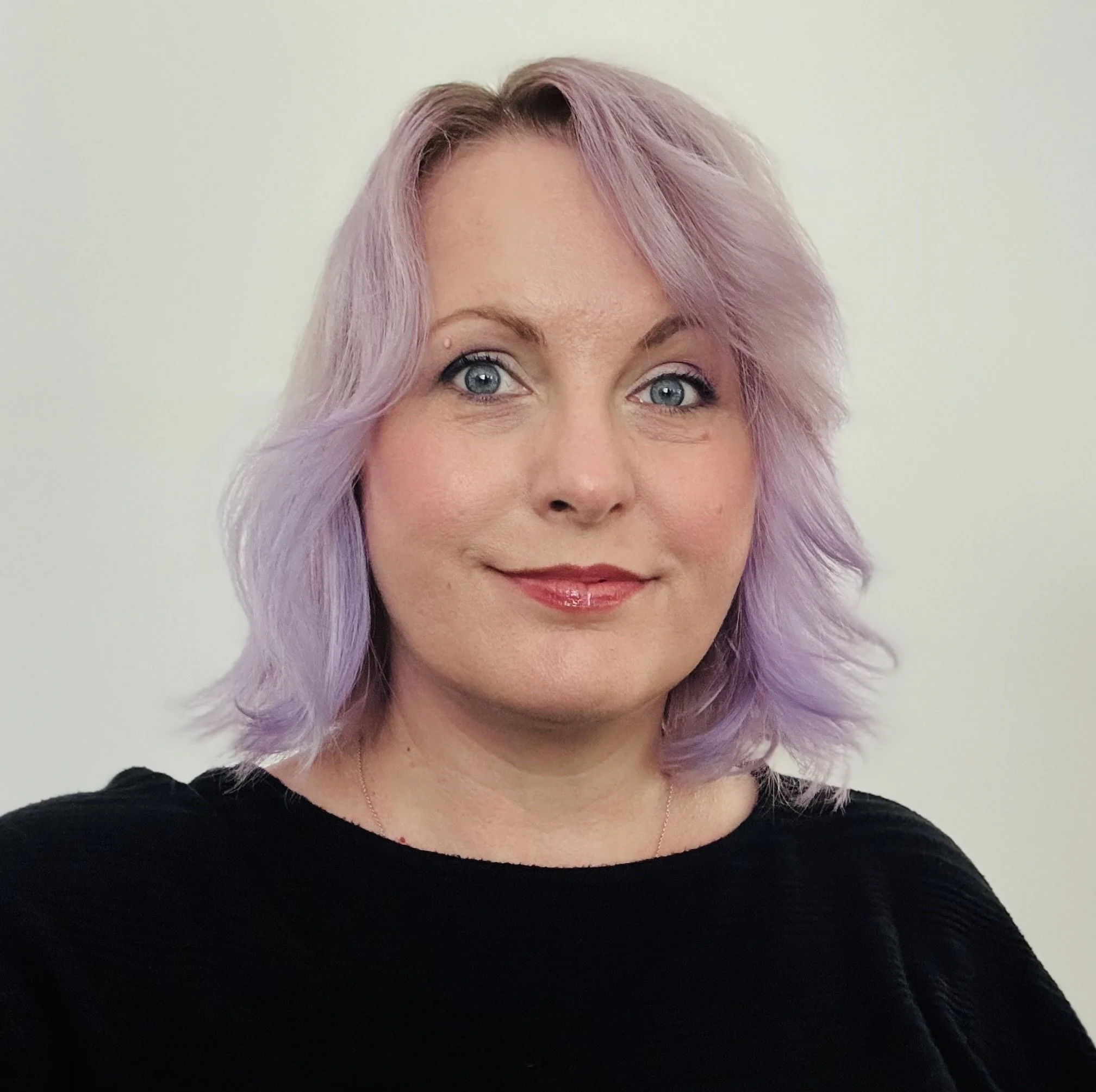 Close-up of a woman's face showing her eyes, pinkish-purple hair, and part of her black top against a plain light-colored background.