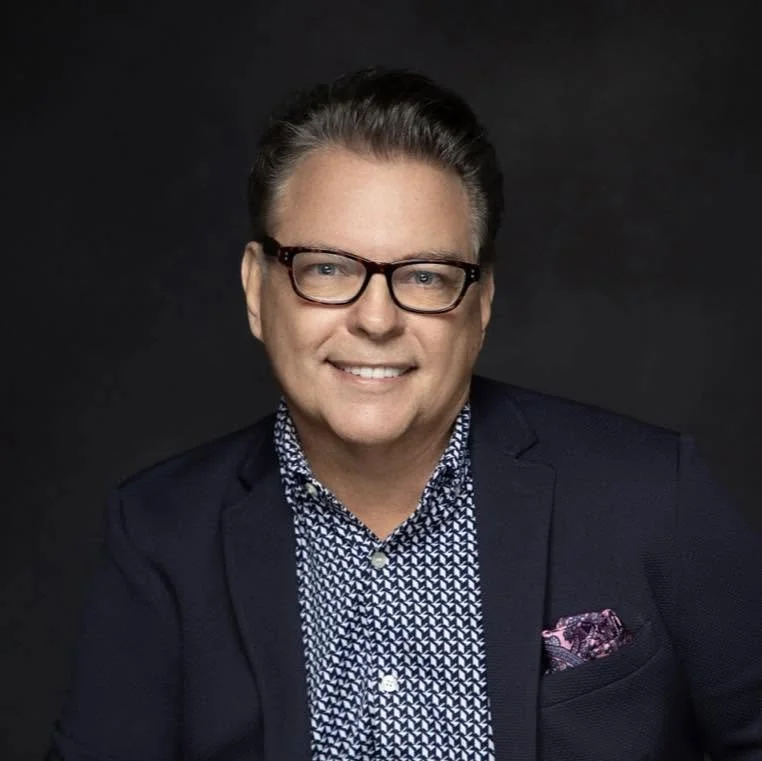 Portrait of a man with glasses, short neatly styled hair, wearing a dark blazer, patterned shirt, and pocket square, against a dark background.
