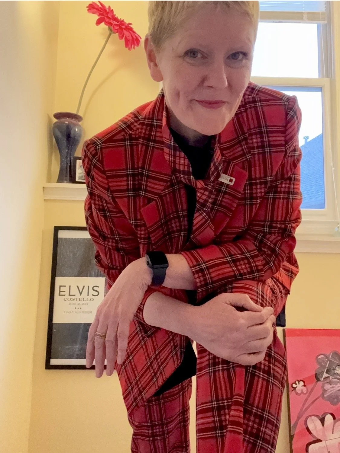 A woman with short red hair wearing a red plaid suit, standing in a room with yellow walls, a window, and framed art, including a poster of Elvis Costello.