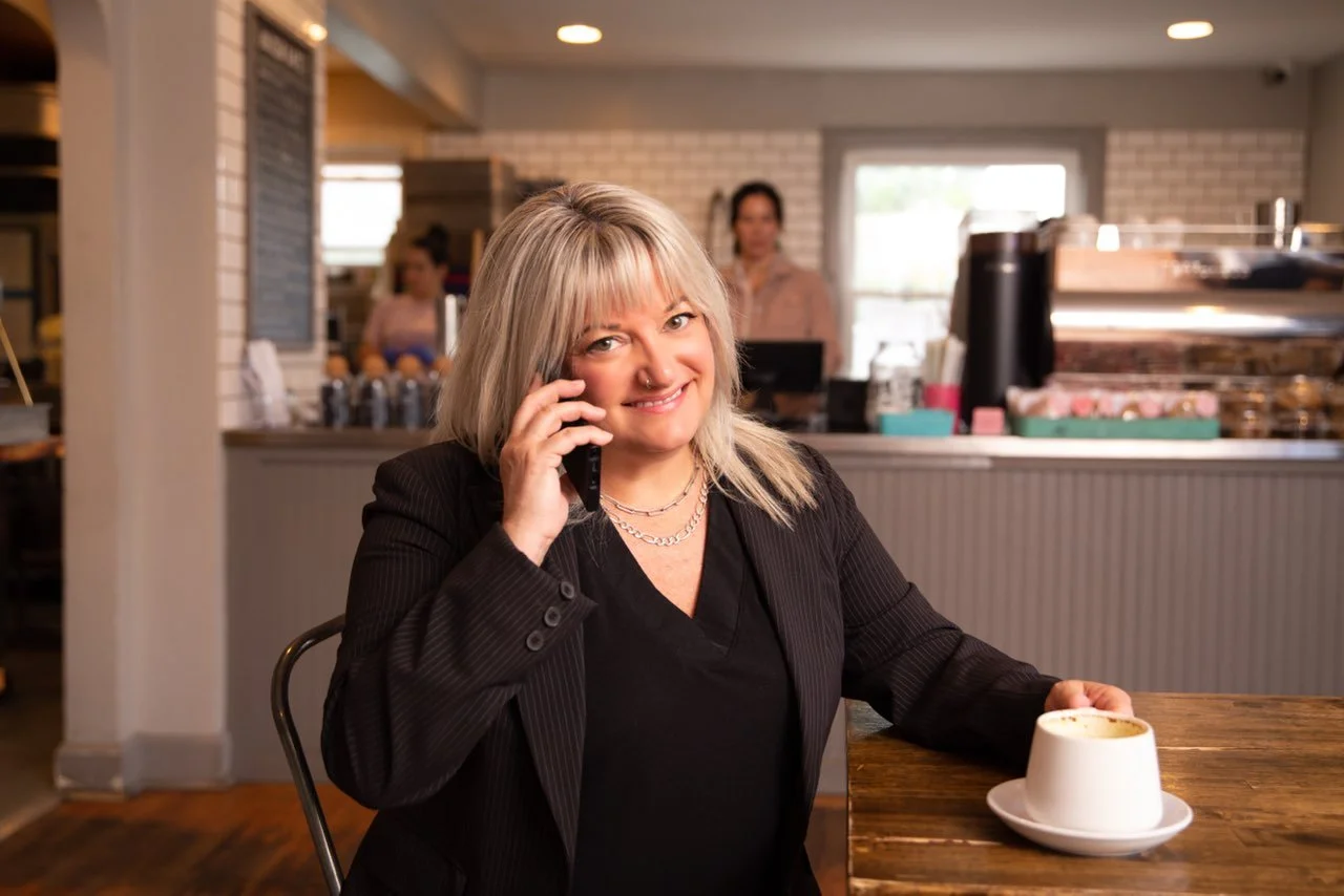 A woman with blonde hair smiling while talking on her phone at a coffee shop, with a cappuccino on the table in front of her.