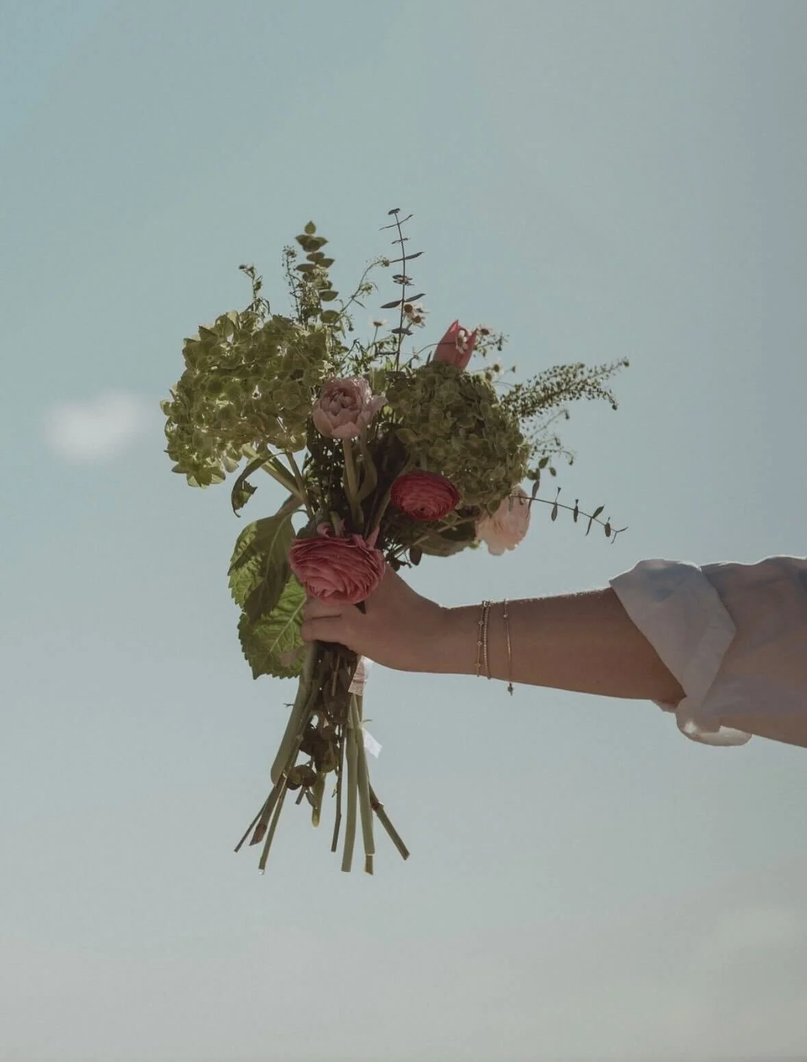 Person holding a bouquet of pink and green flowers against a light blue sky.