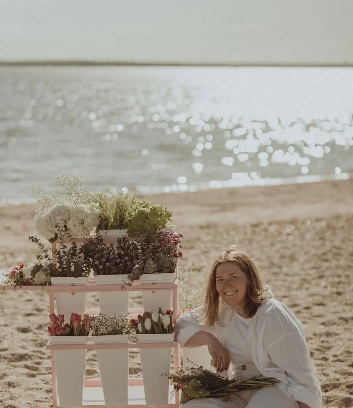 A smiling woman with shoulder-length hair sitting on a beach near a white wooden flower stand with various flowers in pots, with water and the horizon in the background.