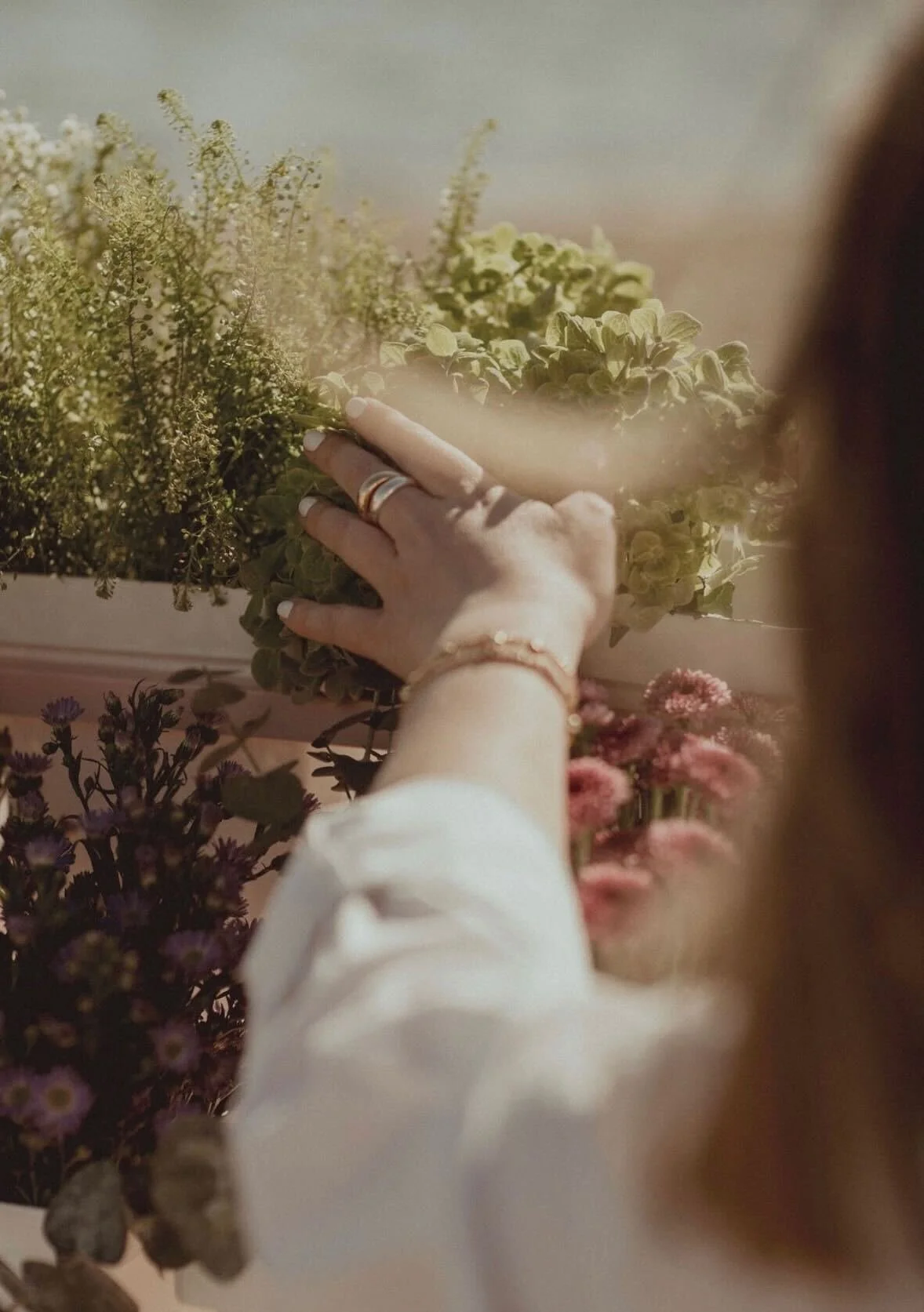 A person with rings on fingers and a bracelet reaches out to touch a bouquet of yellow flowers among other floral arrangements with purple and pink flowers.