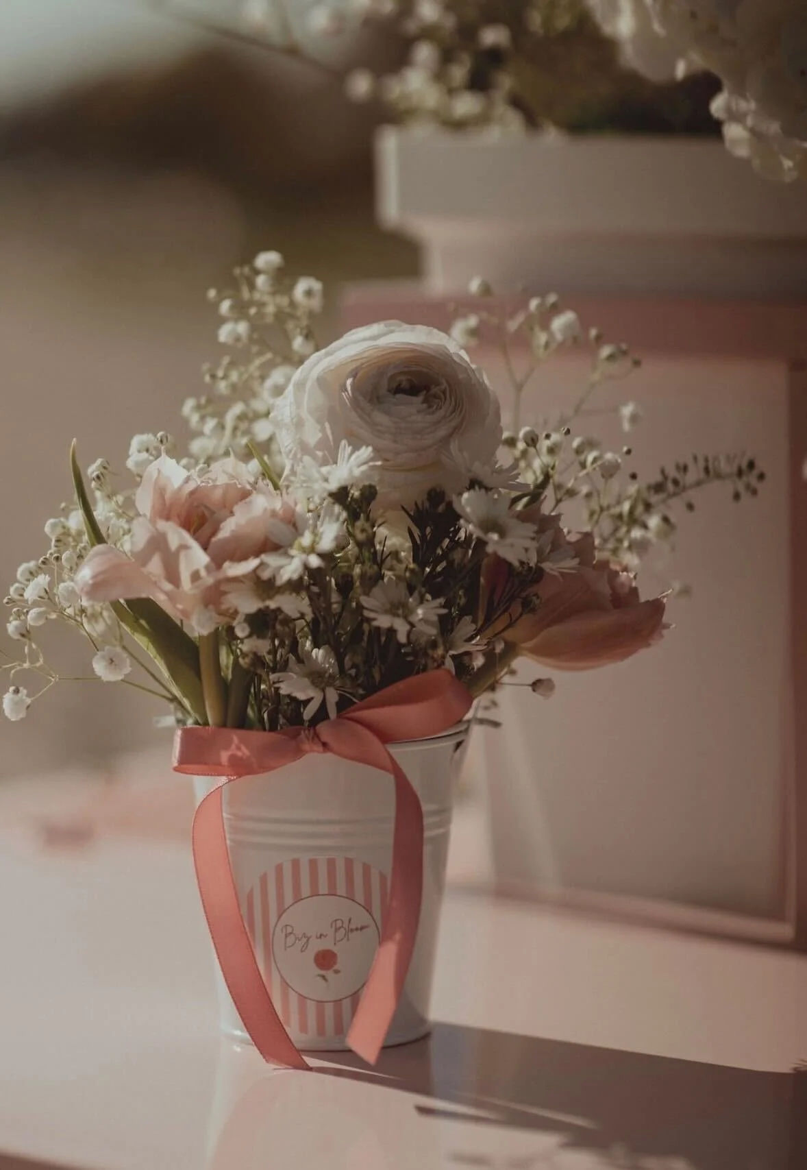 A small bouquet of pink and white flowers in a white cup with a pink ribbon tied around it, labeled "Buy in Bloom".
