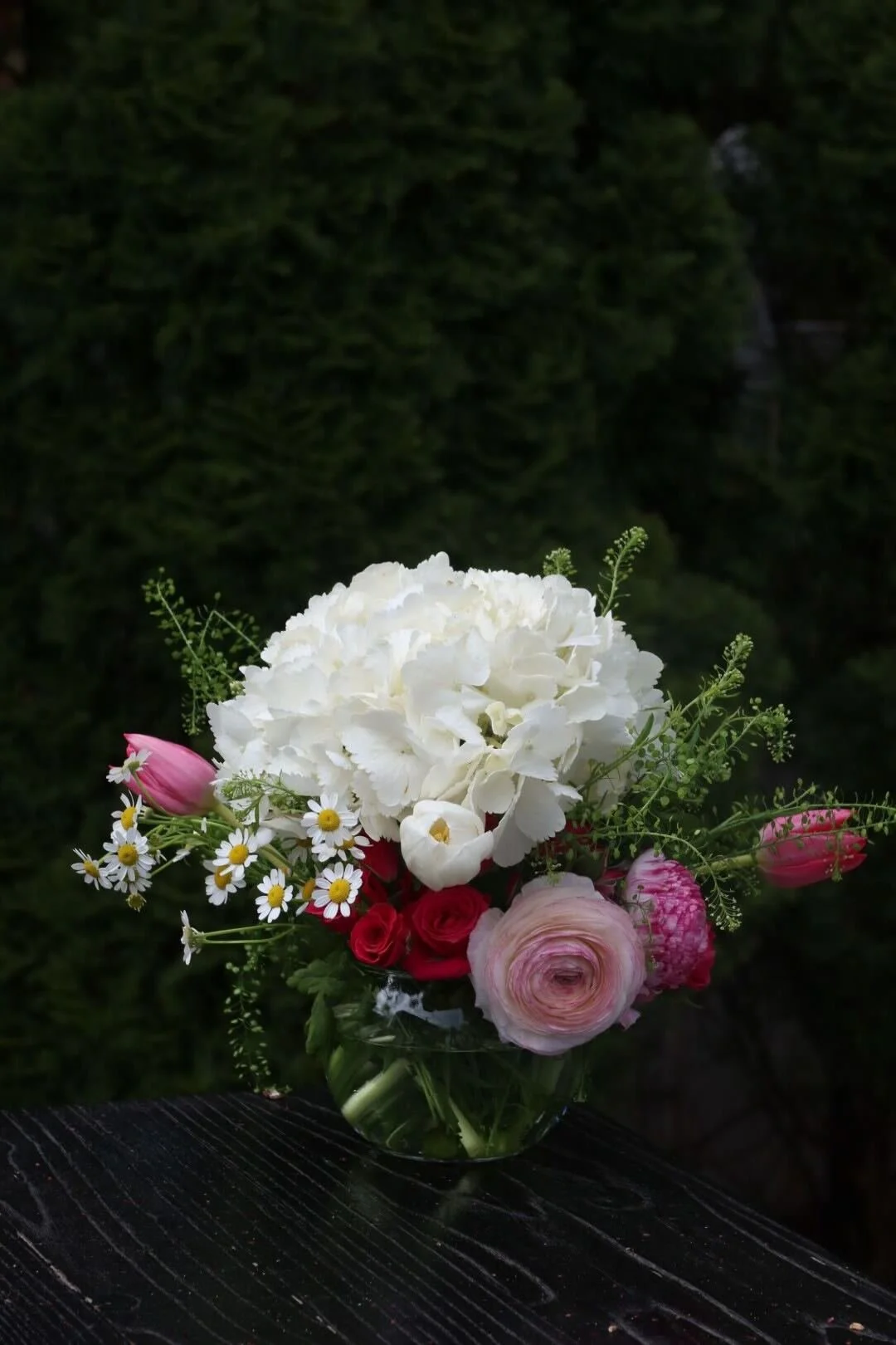 A floral bouquet with white hydrangeas, pink ranunculus and tulips, small daisies, and greenery in a clear glass vase on a black wooden surface against a dark leafy background.