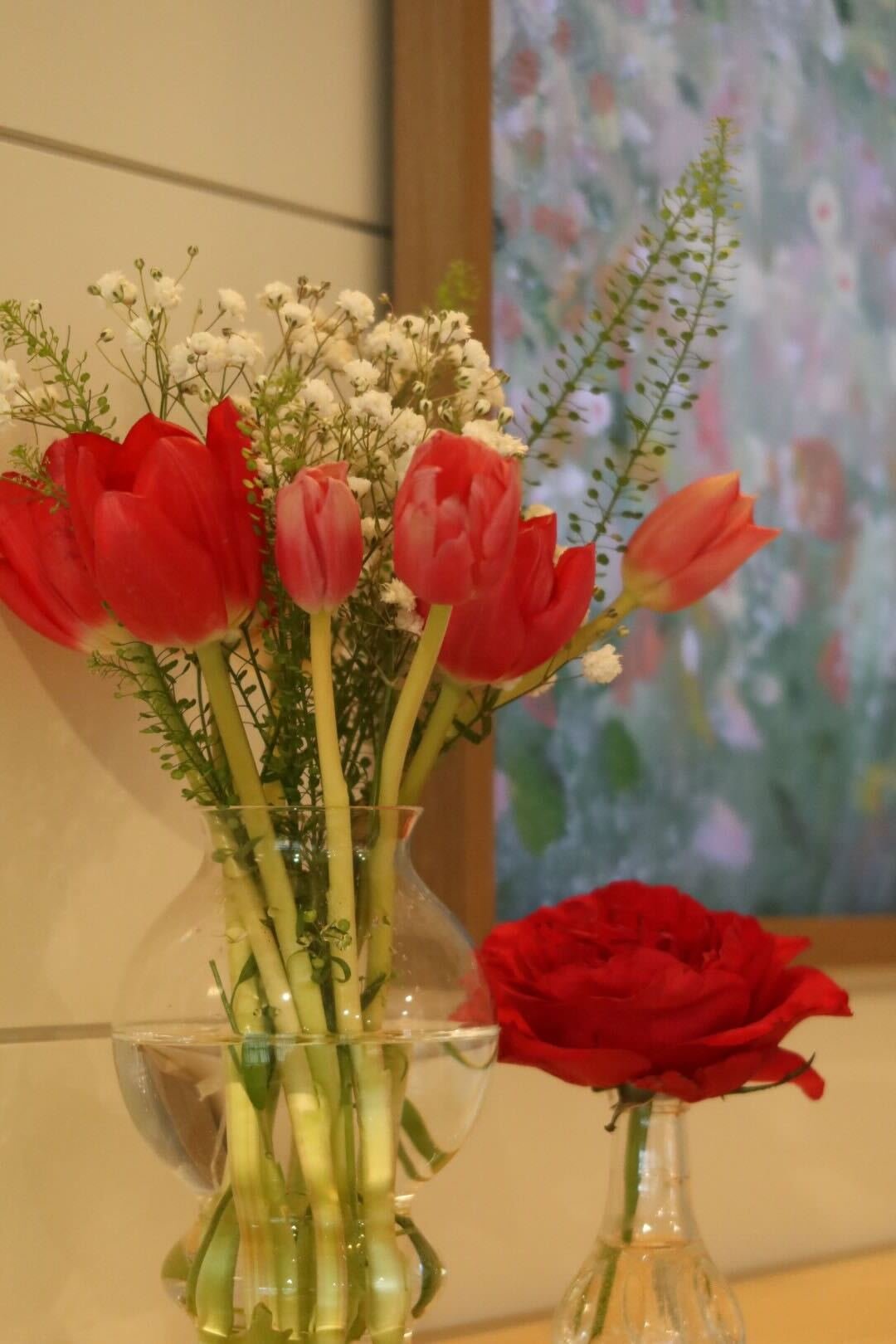 A glass vase with pink tulips, white baby's breath, and greenery, along with a single red rose in a small glass vase, placed on a white surface near a window with a floral curtain.