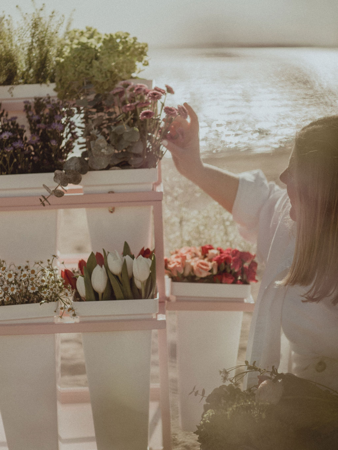 A woman arranging flowers in a flower shop with baskets of various flowers, including tulips, daisies, and roses, near a body of water at sunset.
