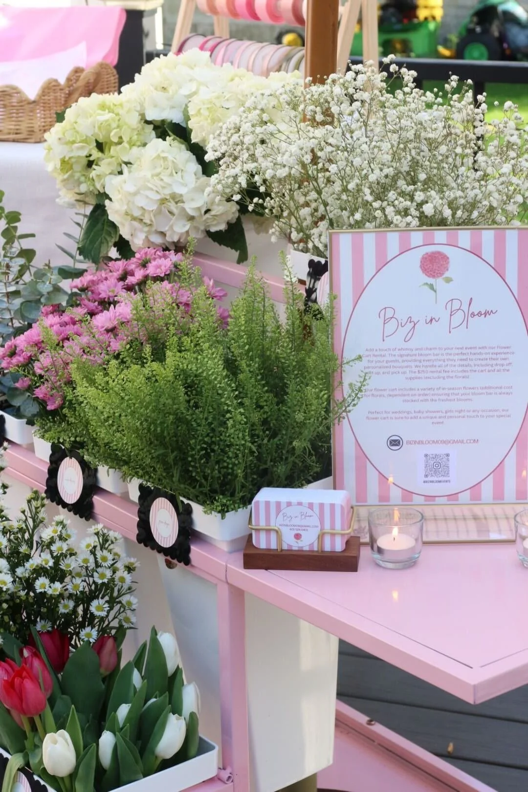 Display of white and pink flowers, including hydrangeas, baby’s breath, and tulips, on a pink table with a sign promoting a flower arrangement service called 'Biz in Bloom'.