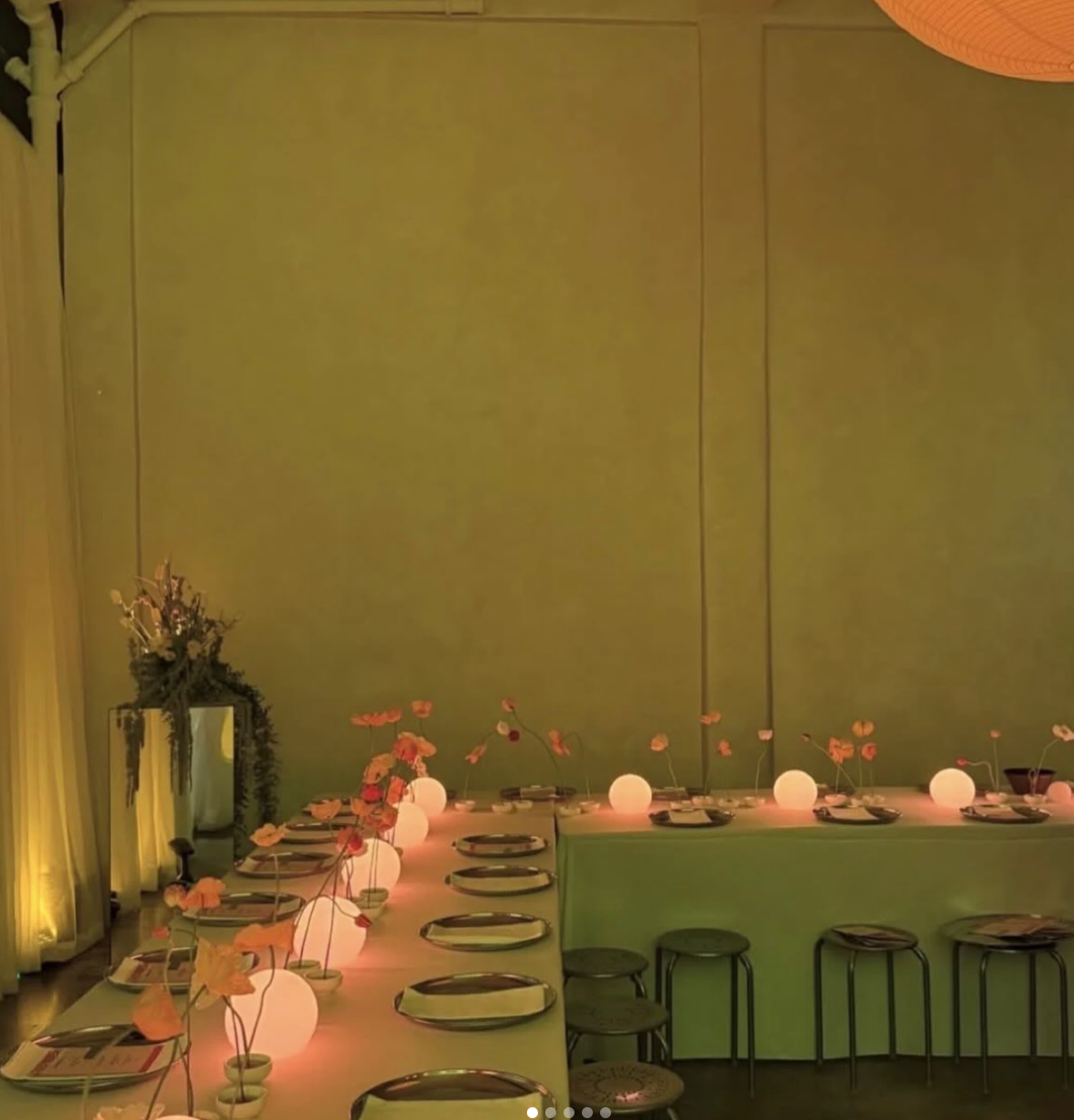 A decorated dining area with a long table set with plates, napkins, and small floral arrangements. The table is illuminated with spherical lamps and surrounded by stools. There is a green wall in the background and a plant arrangement near a mirror on the left.