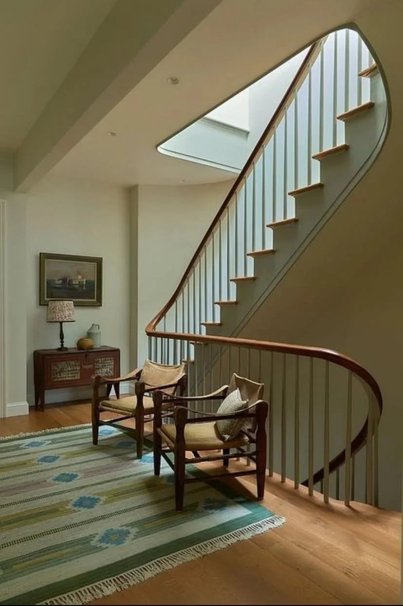 Interior view of a staircase with a wooden handrail, a skylight window above, and cozy seating with two wooden chairs and a patterned cushion on a colorful rug.