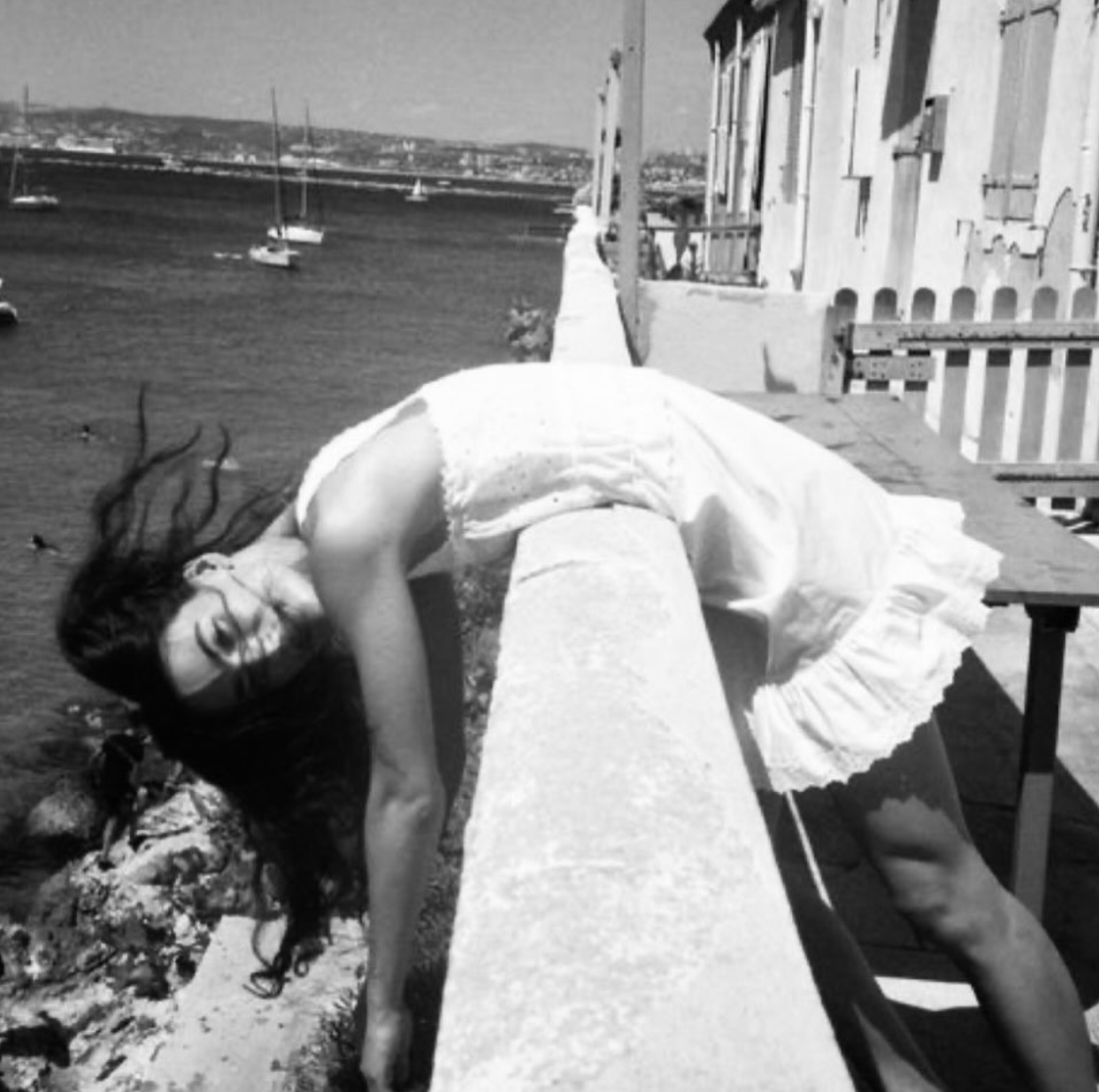 A woman with long hair wearing a light-colored top and skirt leaning over a balcony, with a scenic view of the water and sailboats in the background, on a sunny day.