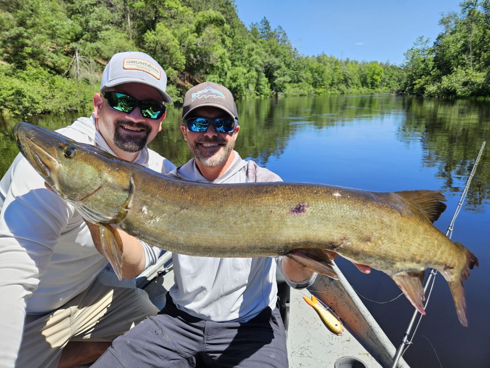 Two men on a boat holding a large fish with a river and green trees in the background.