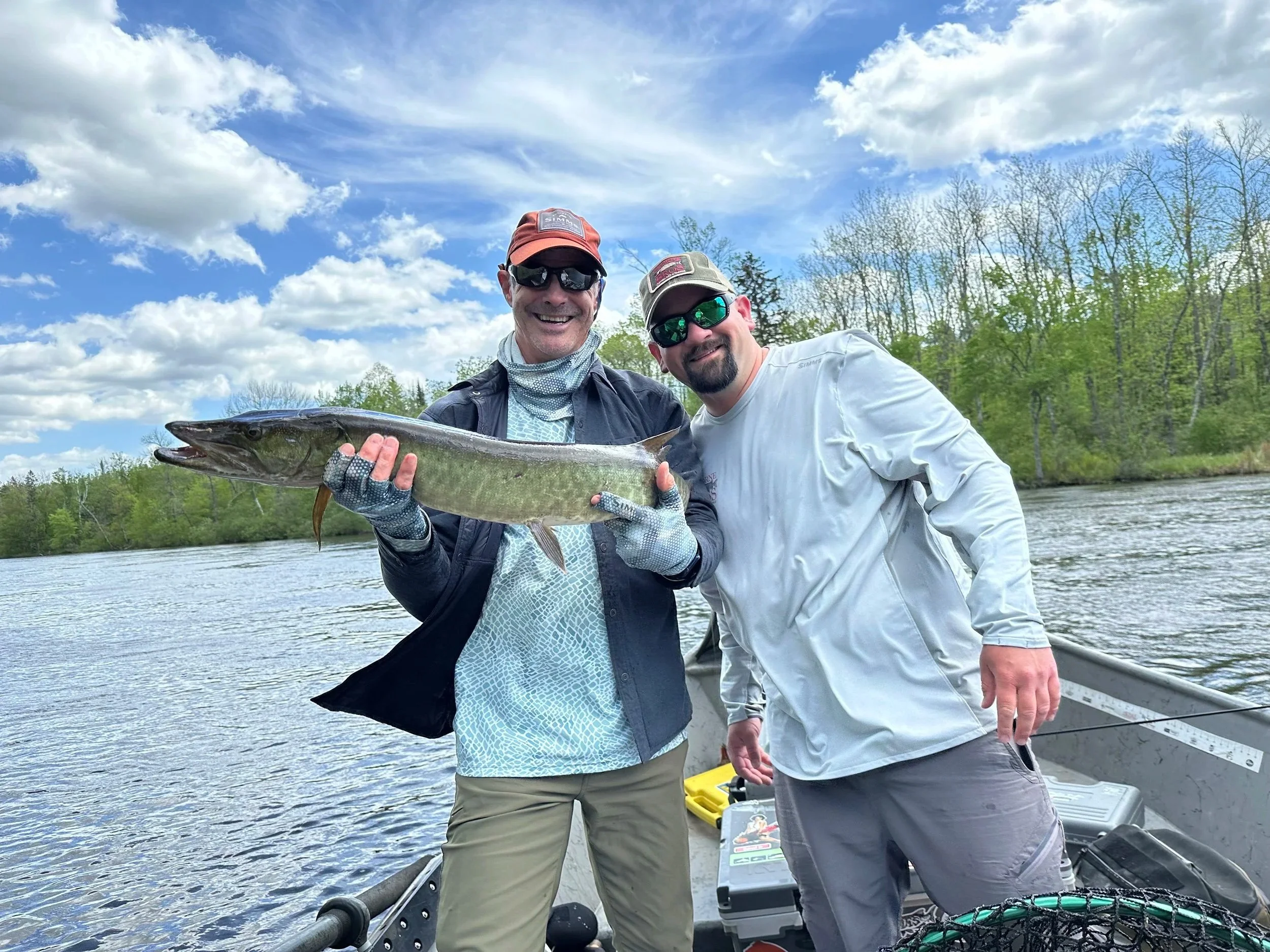 Two men in a boat holding a large fish, against a background of water, trees, and a partly cloudy sky.