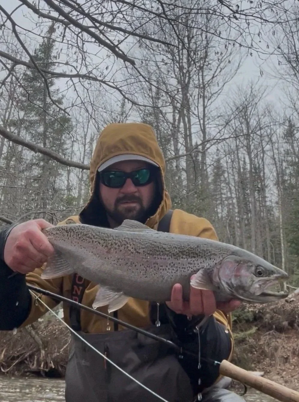 A man wearing a yellow rain jacket, sunglasses, and a hood holding a large rainbow trout fish while sitting in a river surrounded by leafless trees.