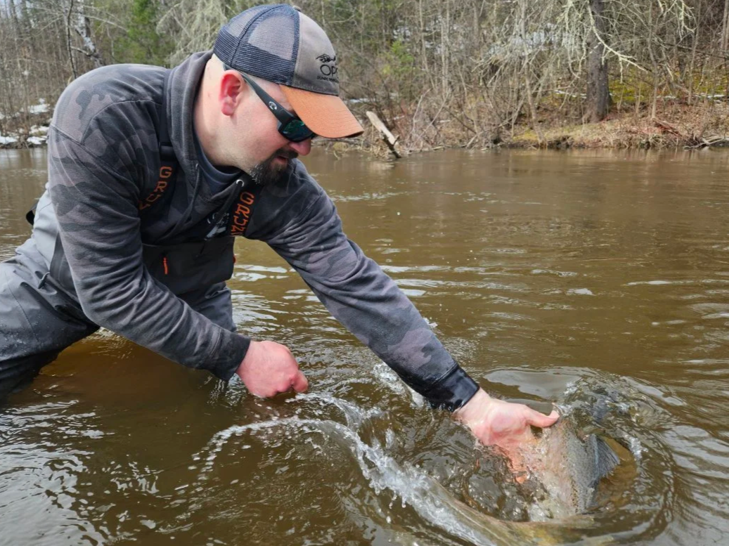 Man wearing a camouflage jacket, baseball cap, and sunglasses leaning into a river, holding a fish with his hands.