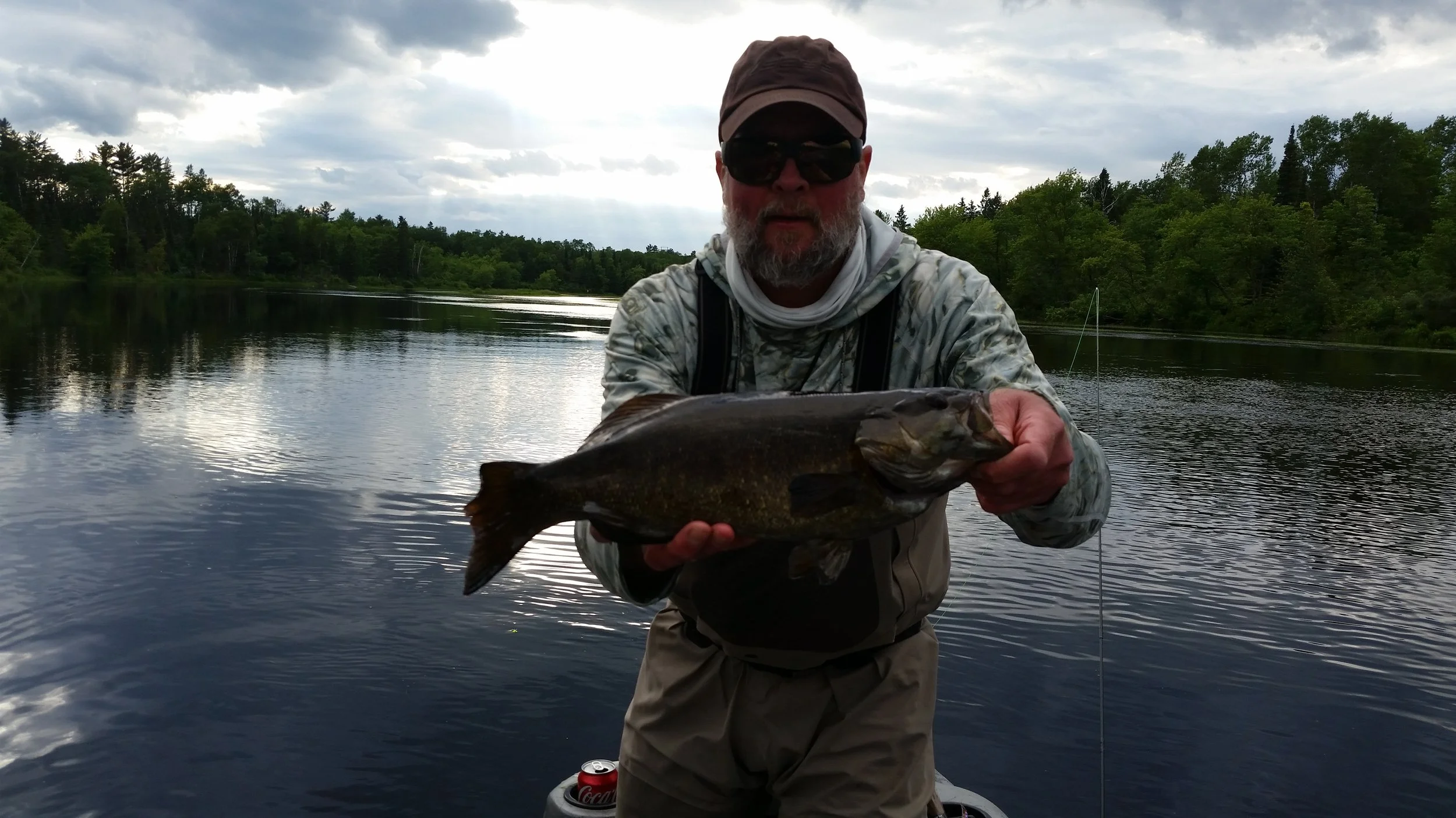Man holding a fish he caught while standing in a boat on a river or lake, with green trees and cloudy sky in the background.