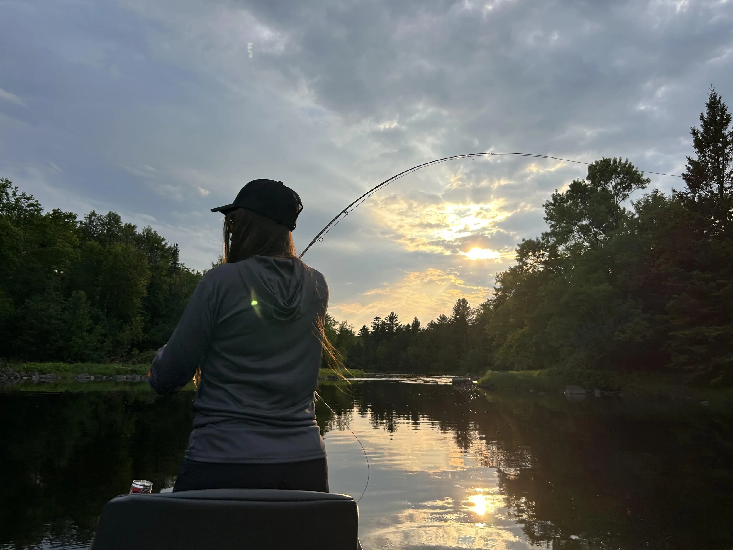 Person fishing in a river at sunset with trees and clouds in the background.