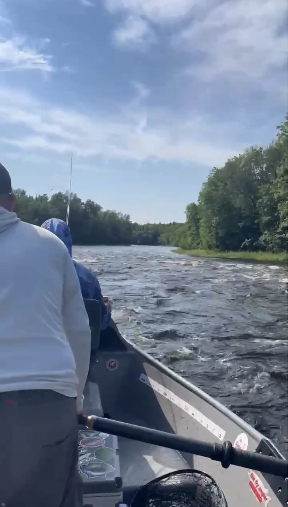 View from a boat on a river, with people sitting and rowing. Two individuals wear hoodies, and much of the boat's interior and a bicycle wheel are visible. The river is surrounded by green trees, and the sky is partly cloudy with blue patches.