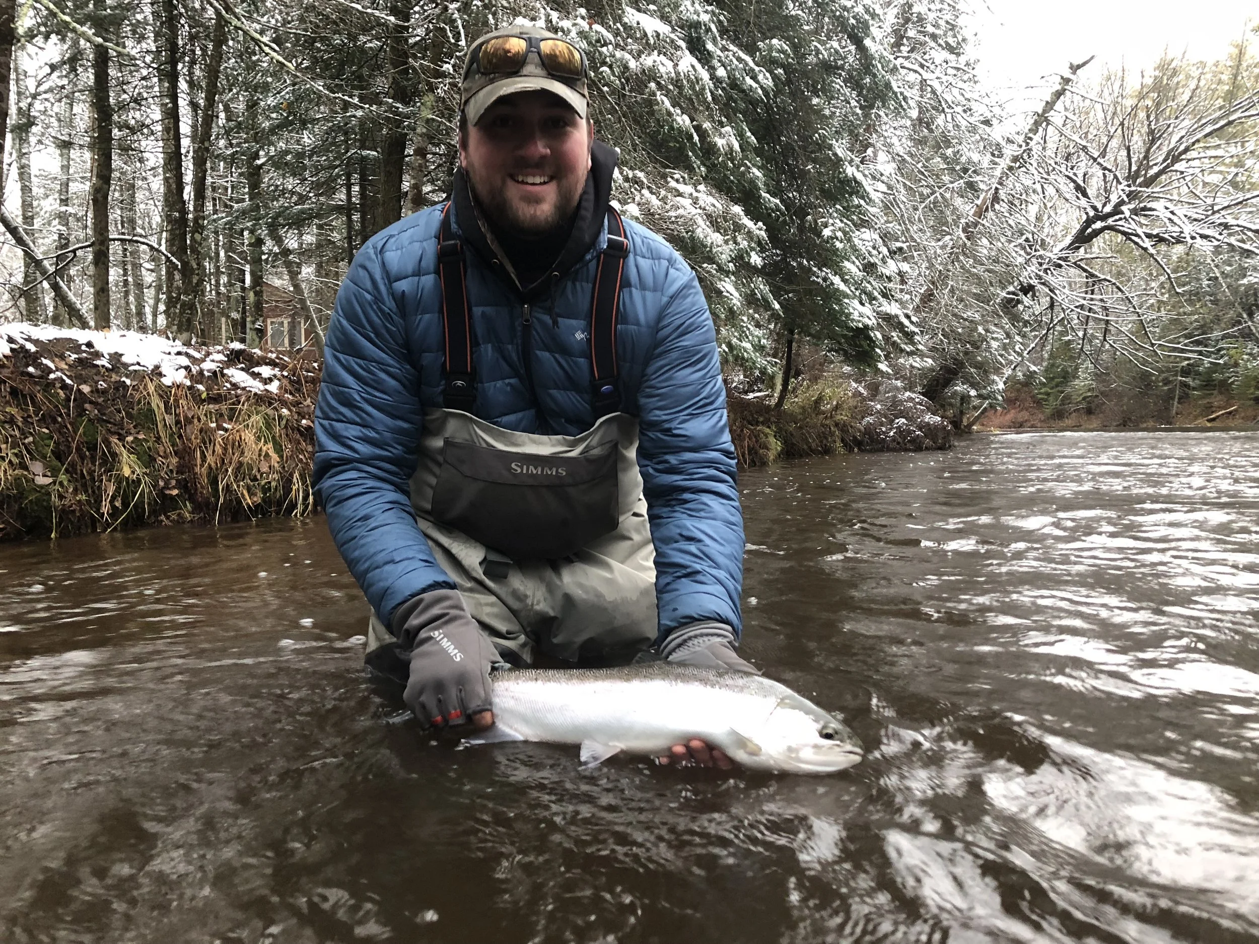 A man smiling and kneeling in a river, holding a fish, with a snowy forest in the background.