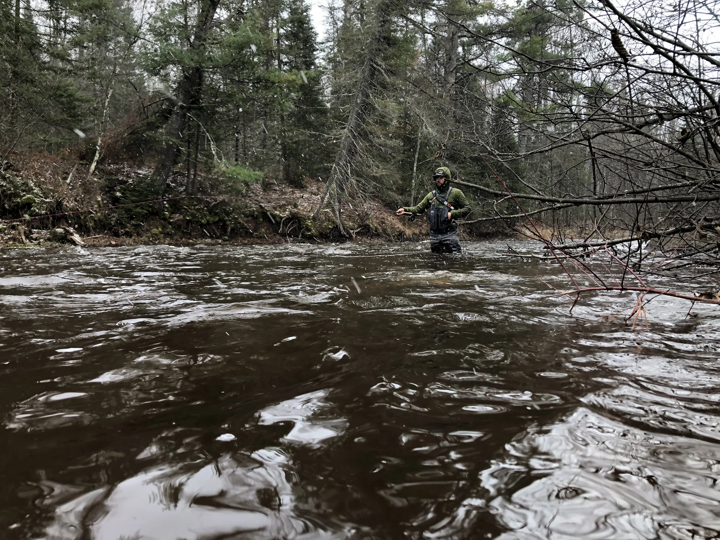 A man standing in a river surrounded by trees, wearing a green jacket, helmet, and fishing gear, fishing with a rod.