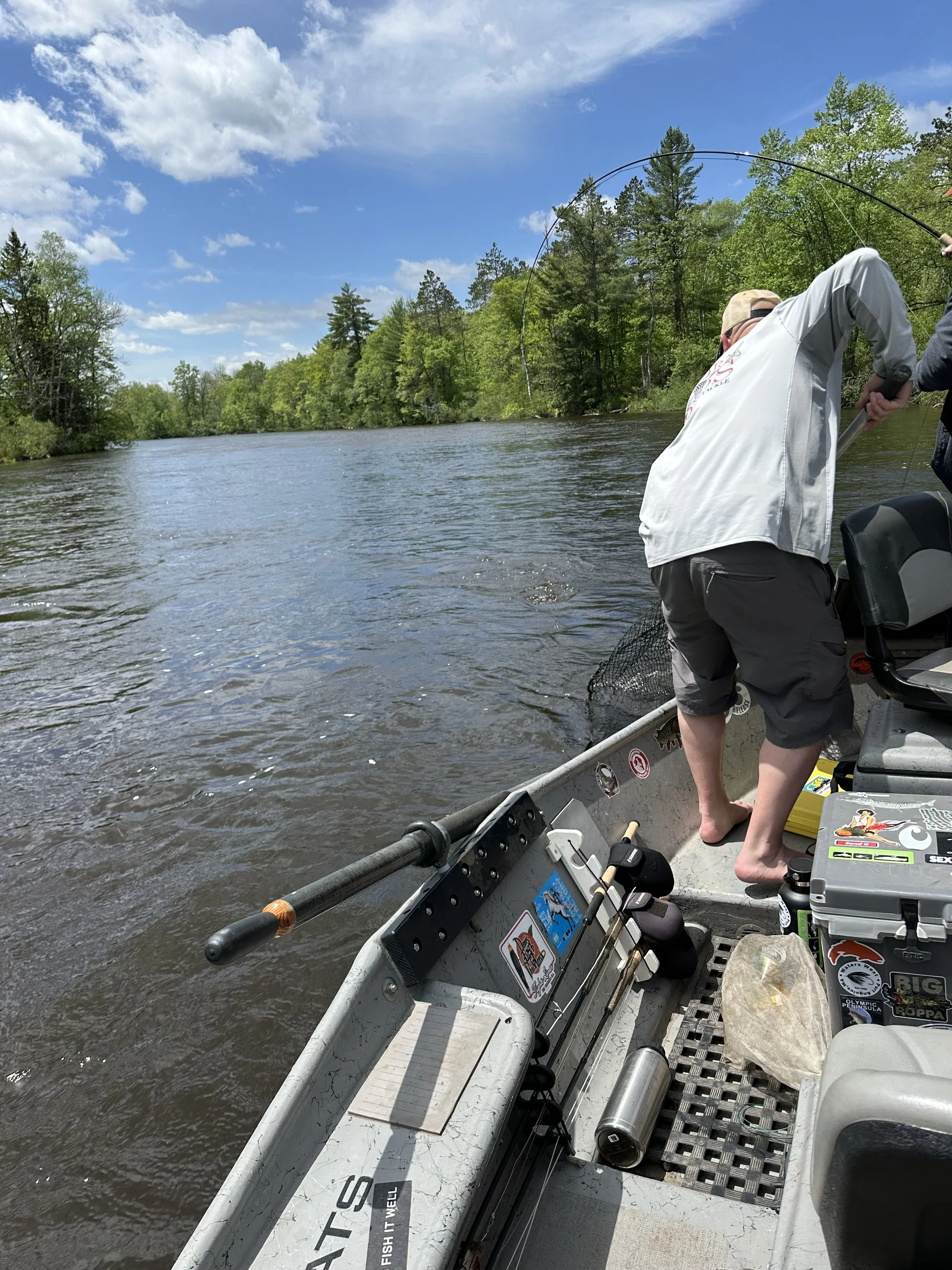 A man fishing from a small boat on a river surrounded by green trees under a partly cloudy sky.