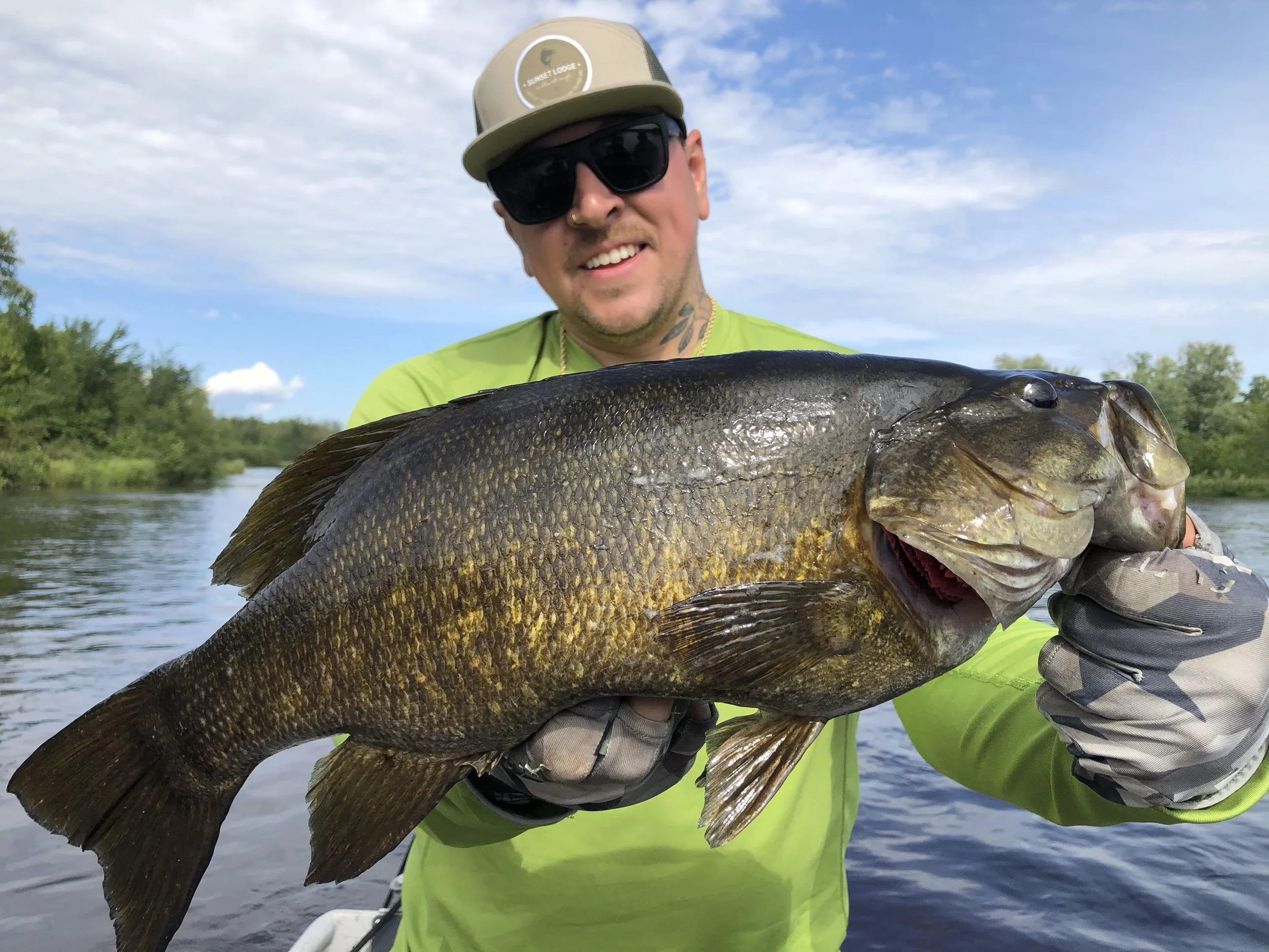 A man with a beard and tattoos holding a large fish outdoors, wearing sunglasses, a beige cap, and a neon green shirt, near a river with a partly cloudy sky.