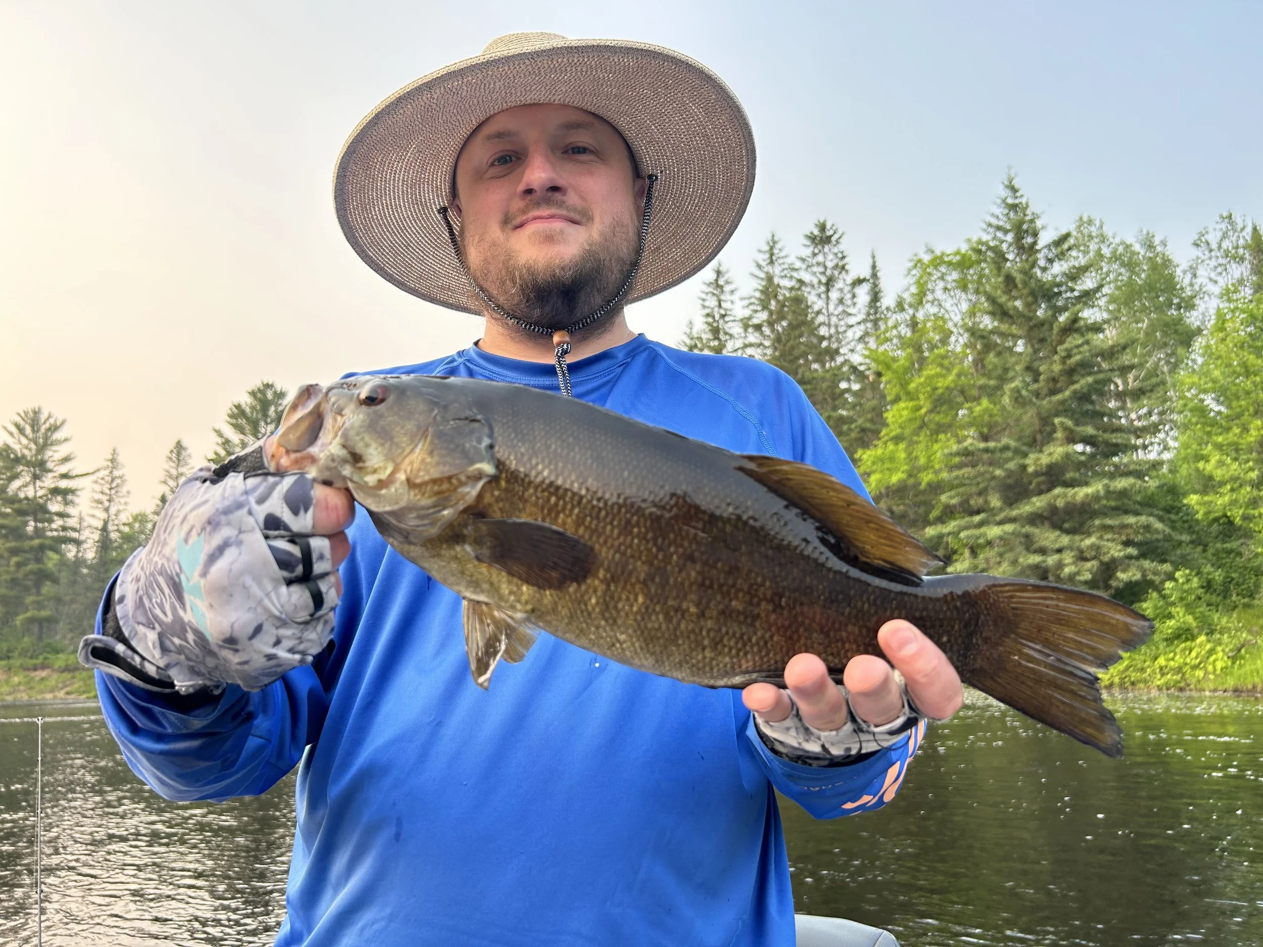 A man wearing a large sun hat and a blue long-sleeve shirt is holding a large fish, smiling, outdoors near a river with trees and clear sky in the background.