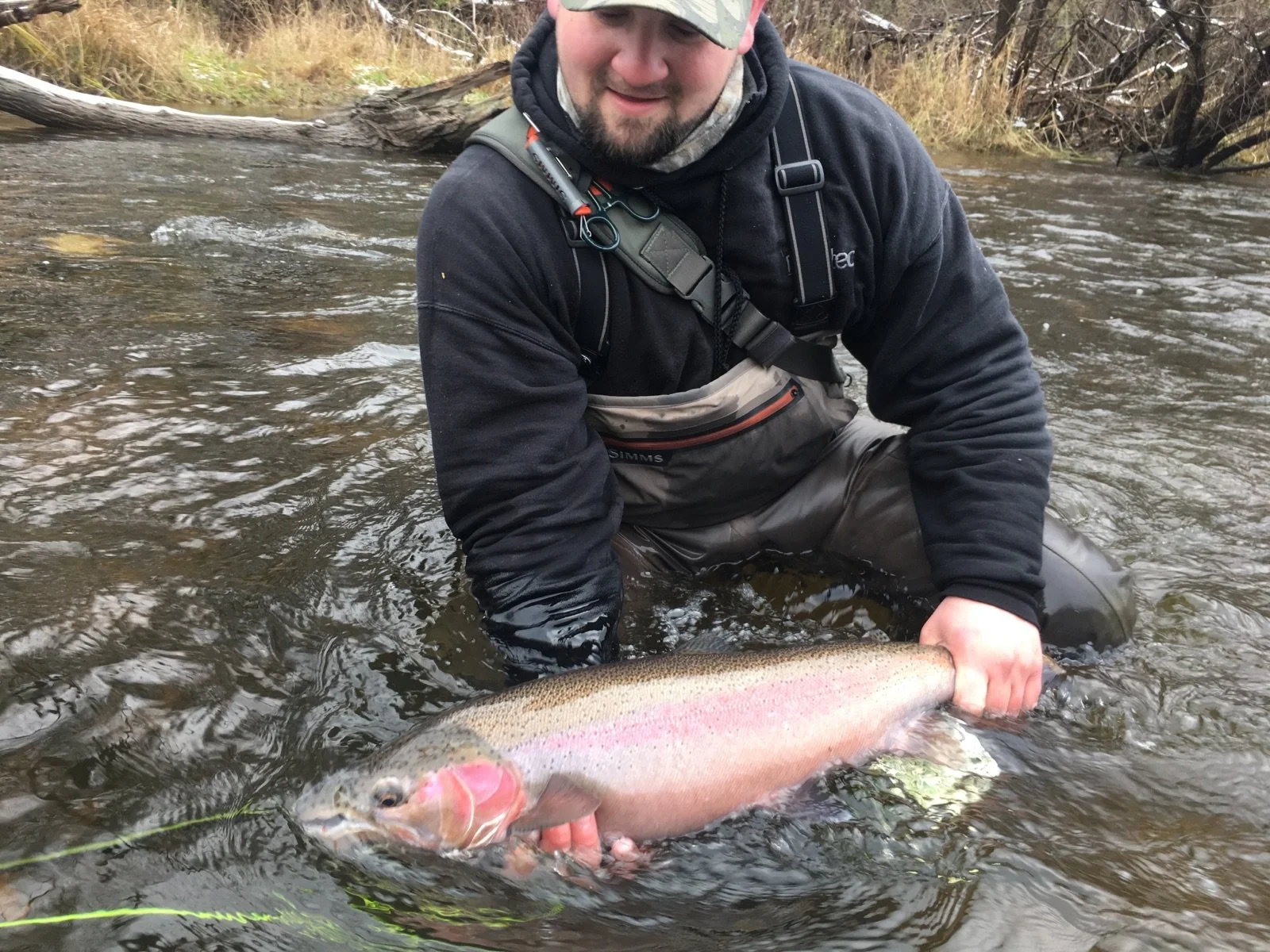 A man in outdoor gear kneeling in a river, holding a large rainbow trout he caught.