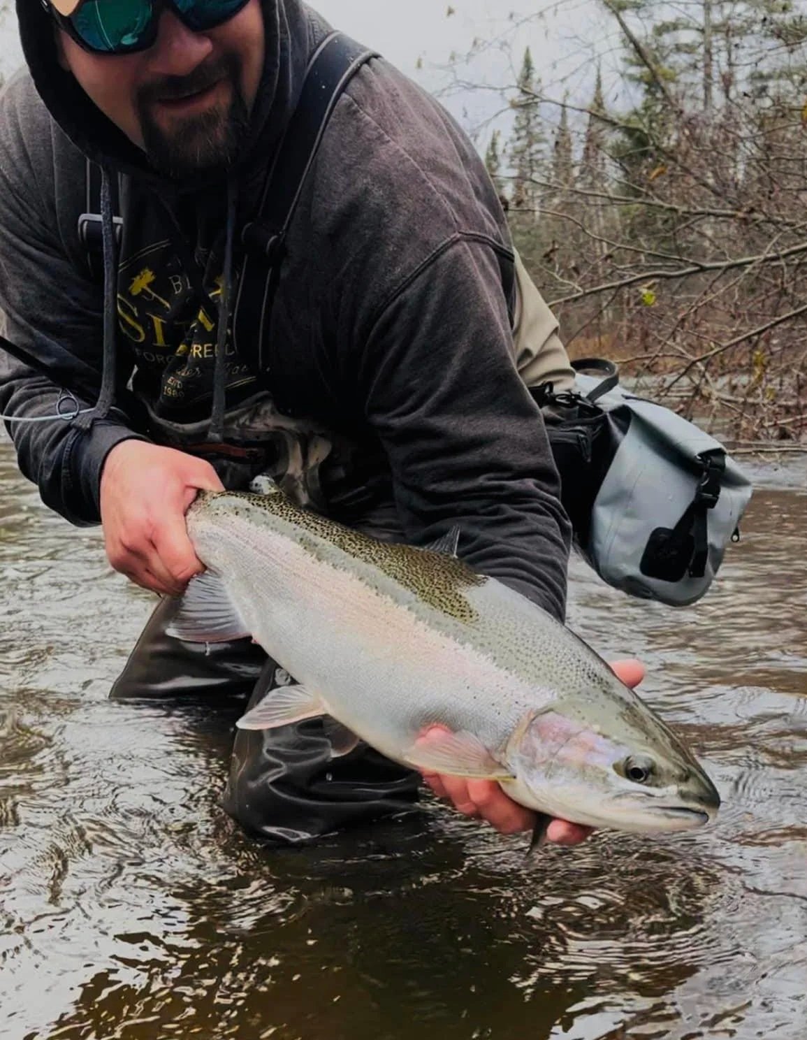 A man is kneeling in a shallow river, holding a large fish, likely a rainbow trout, with both hands. He is wearing dark sunglasses, a gray long-sleeve shirt, and a black jacket. The background features leafless trees and water.