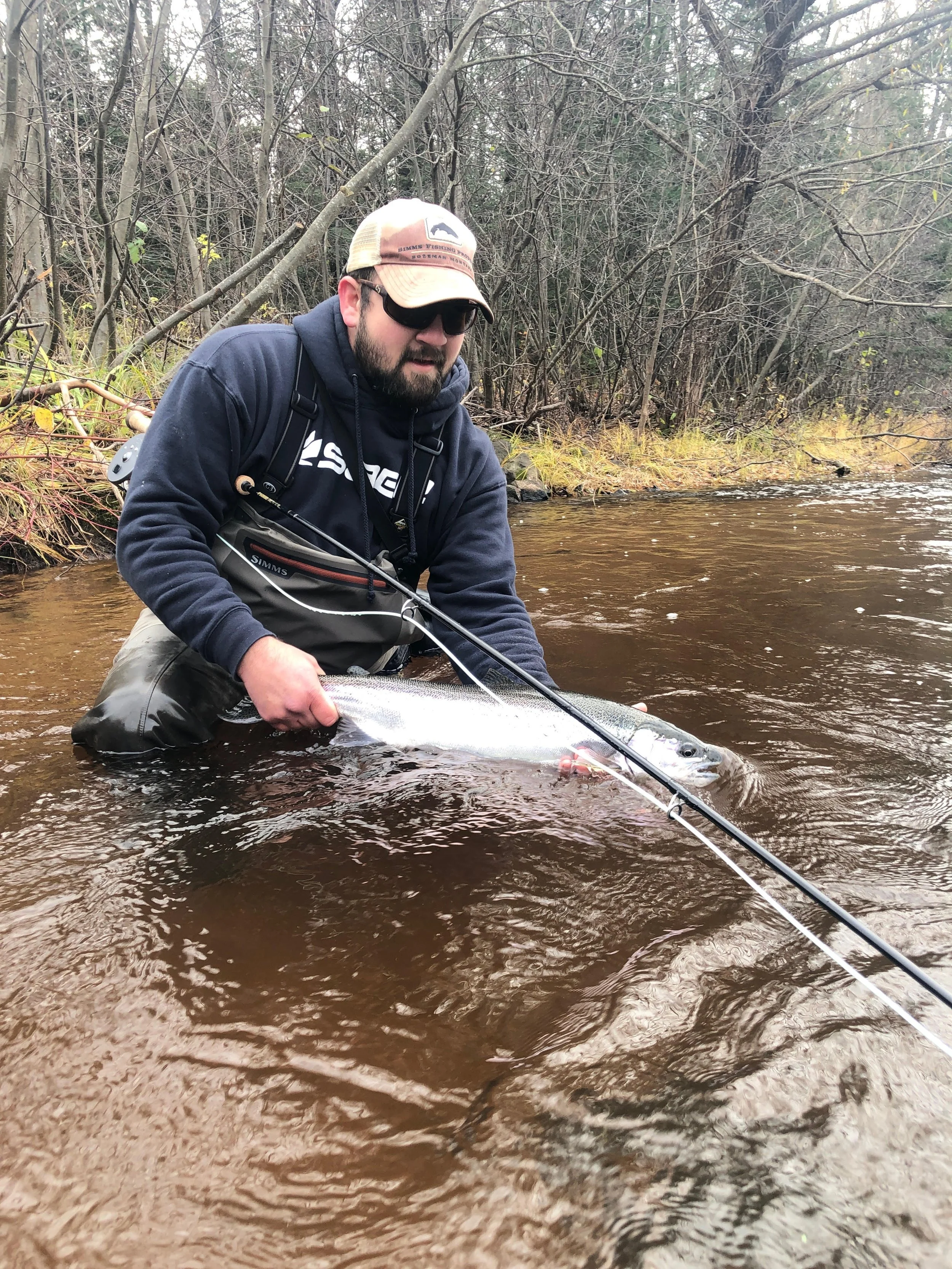 A man in waders and a hoodie, wearing sunglasses and a cap, is kneeling in a stream holding a large fish, likely a salmon or trout, with fishing gear nearby.