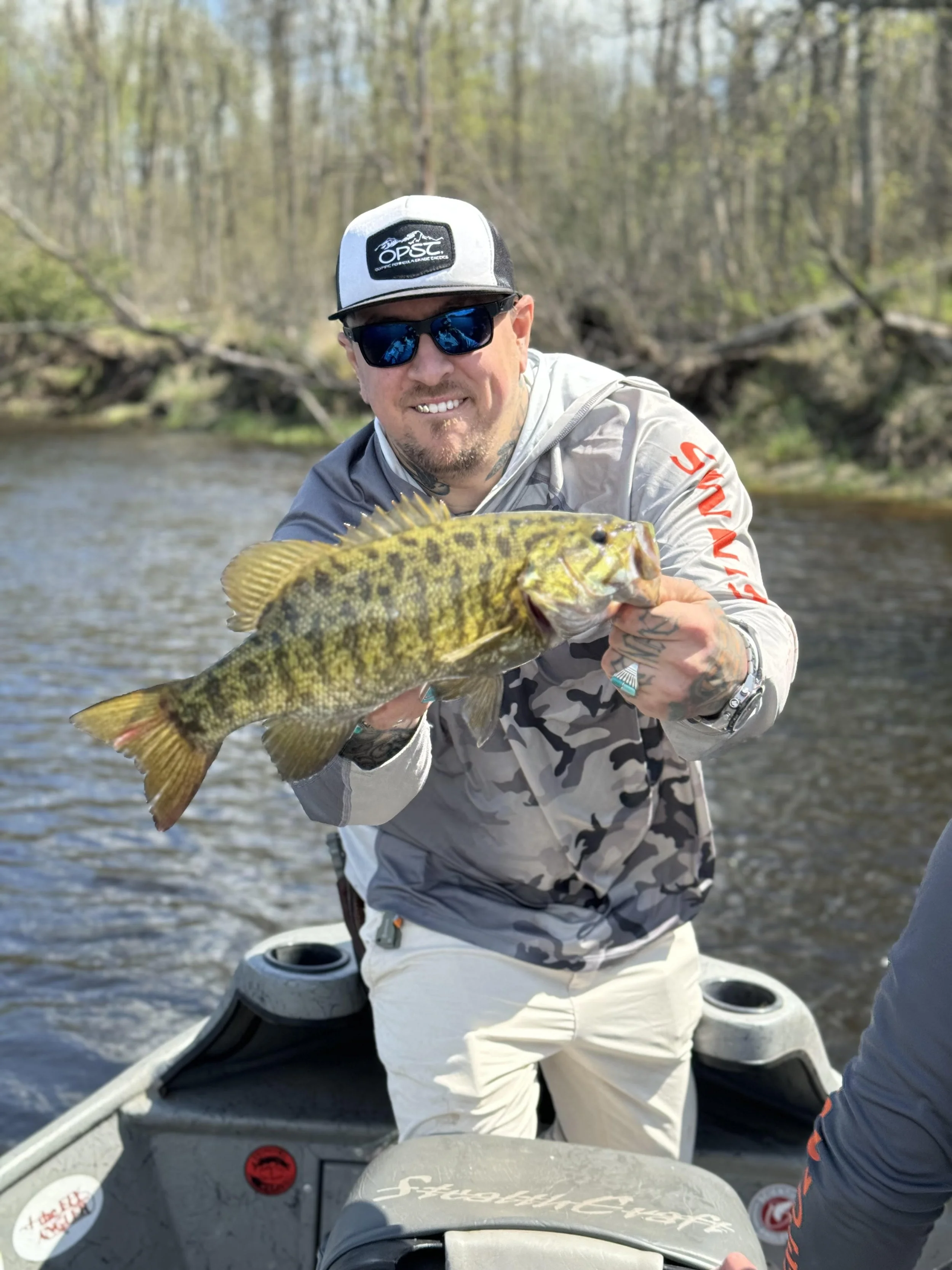 Man holding a yellow and green fish while on a boat in a river, with trees in the background.