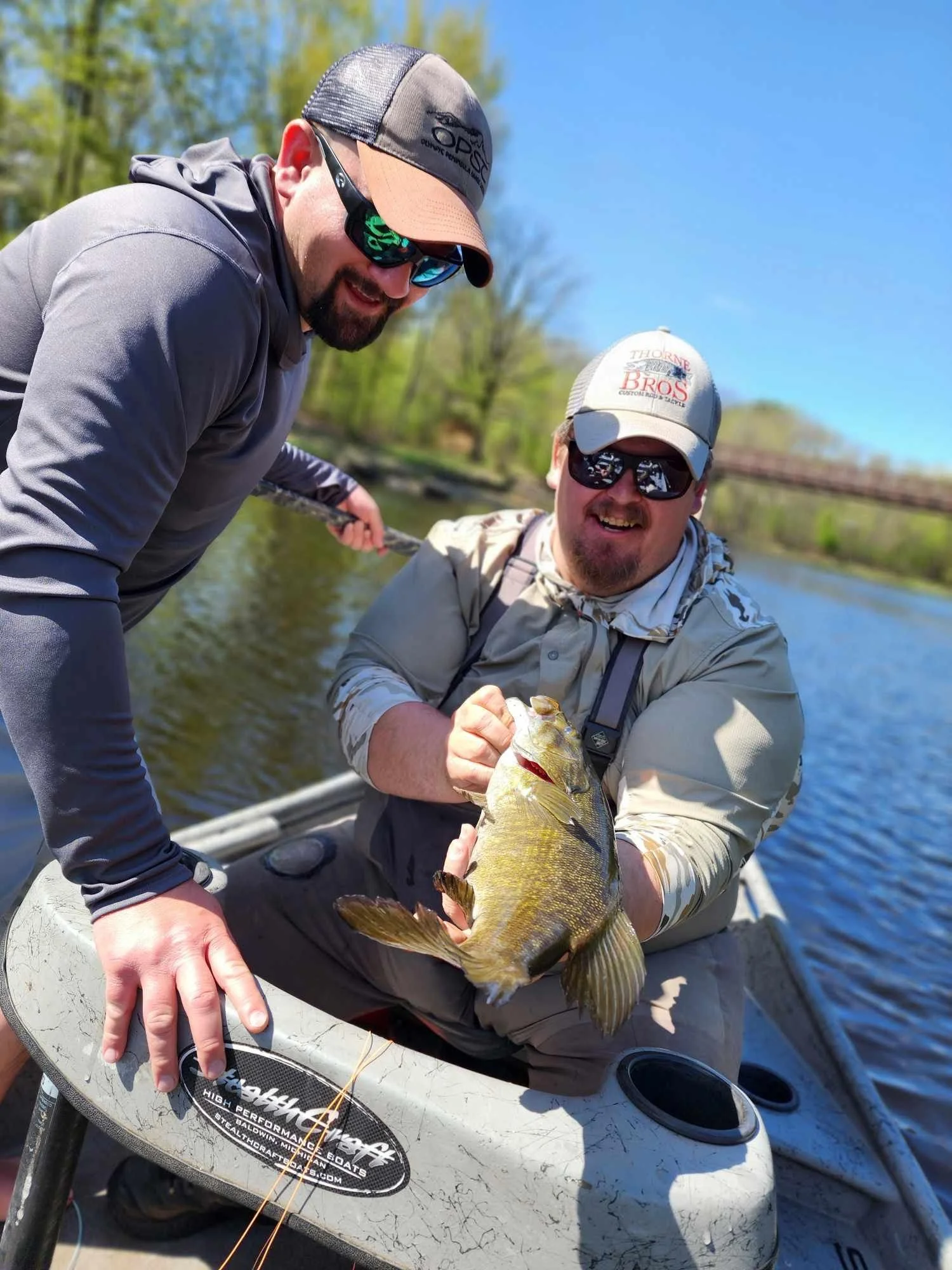 Two men on a boat, one holding a fish, with trees and water in the background, during a sunny day.