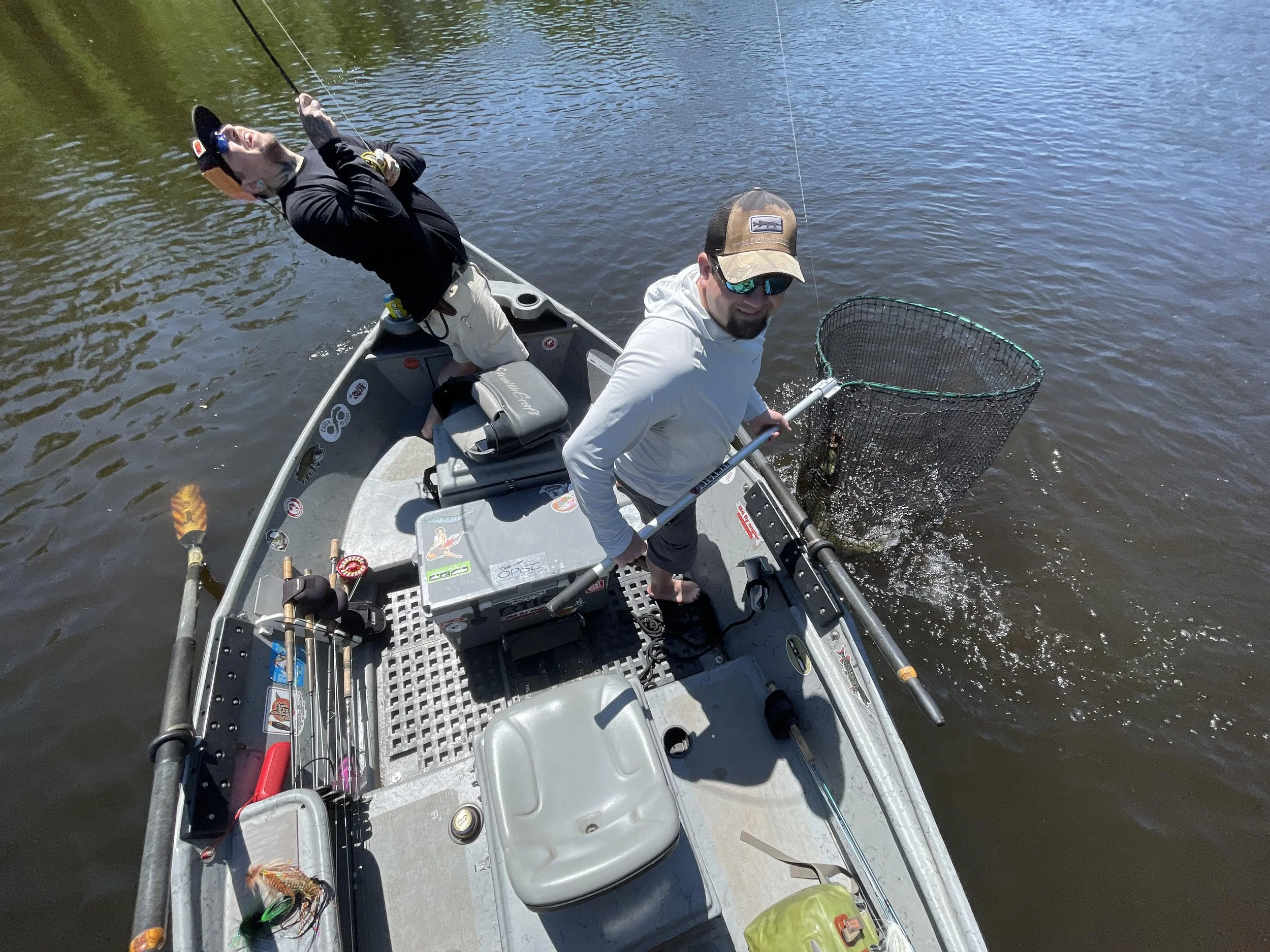 Two men are on a small fishing boat on a river, one holding a fishing net with a fish inside, the other sitting and smiling, wearing sunglasses and a baseball cap. The boat has fishing gear and stickers on it.