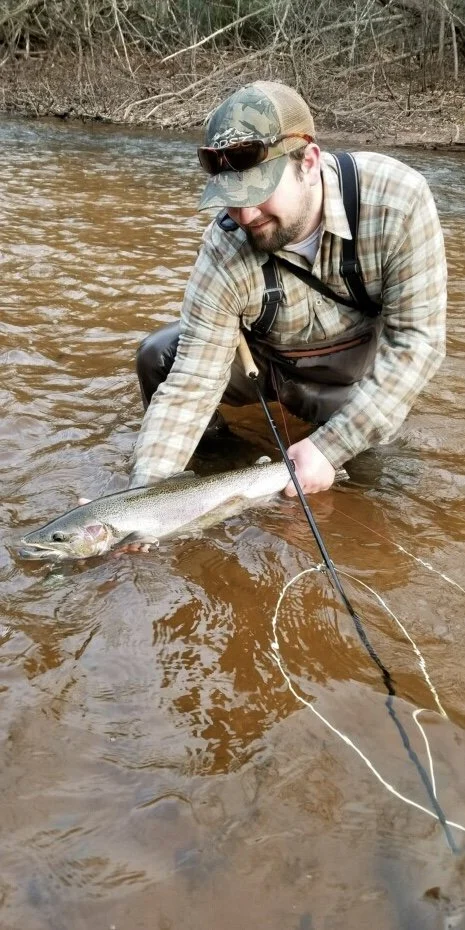 Man in plaid shirt and camouflage cap catching a fish in a river, holding it with both hands while kneeling in the water.