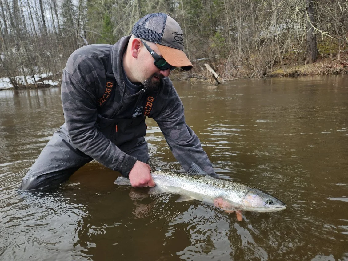 Man wearing sunglasses, a hat, and outdoor gear holds a large fish in a shallow river.