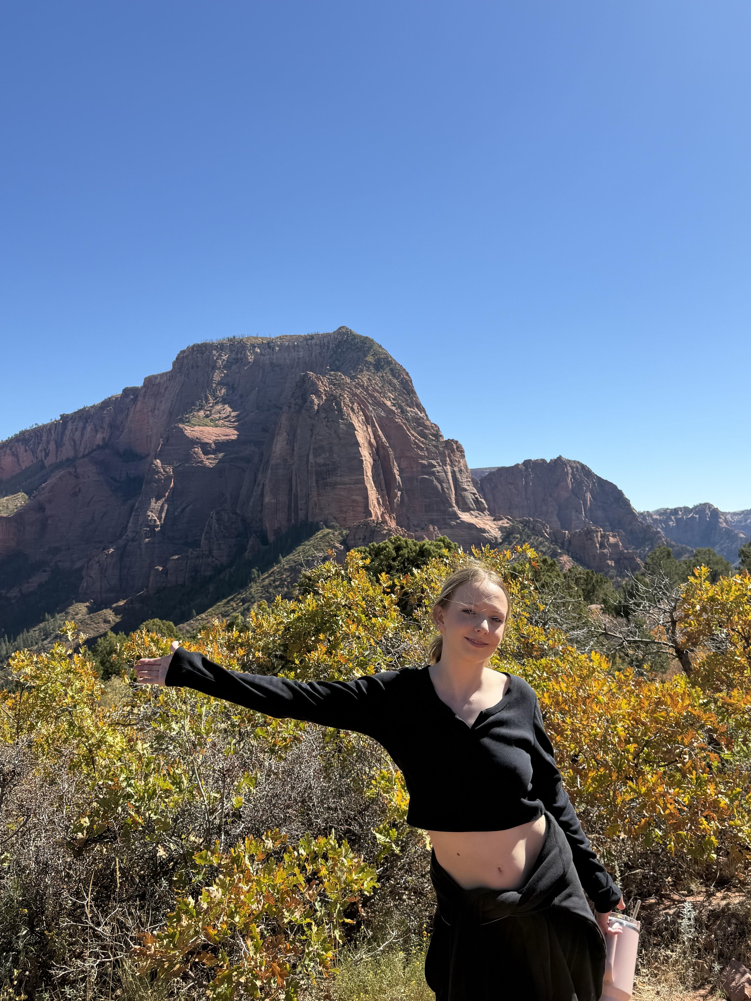 A young woman in black clothing stands with her right arm extended in front of a rocky mountain and autumn foliage, smiling at the camera in a sunny outdoor setting.