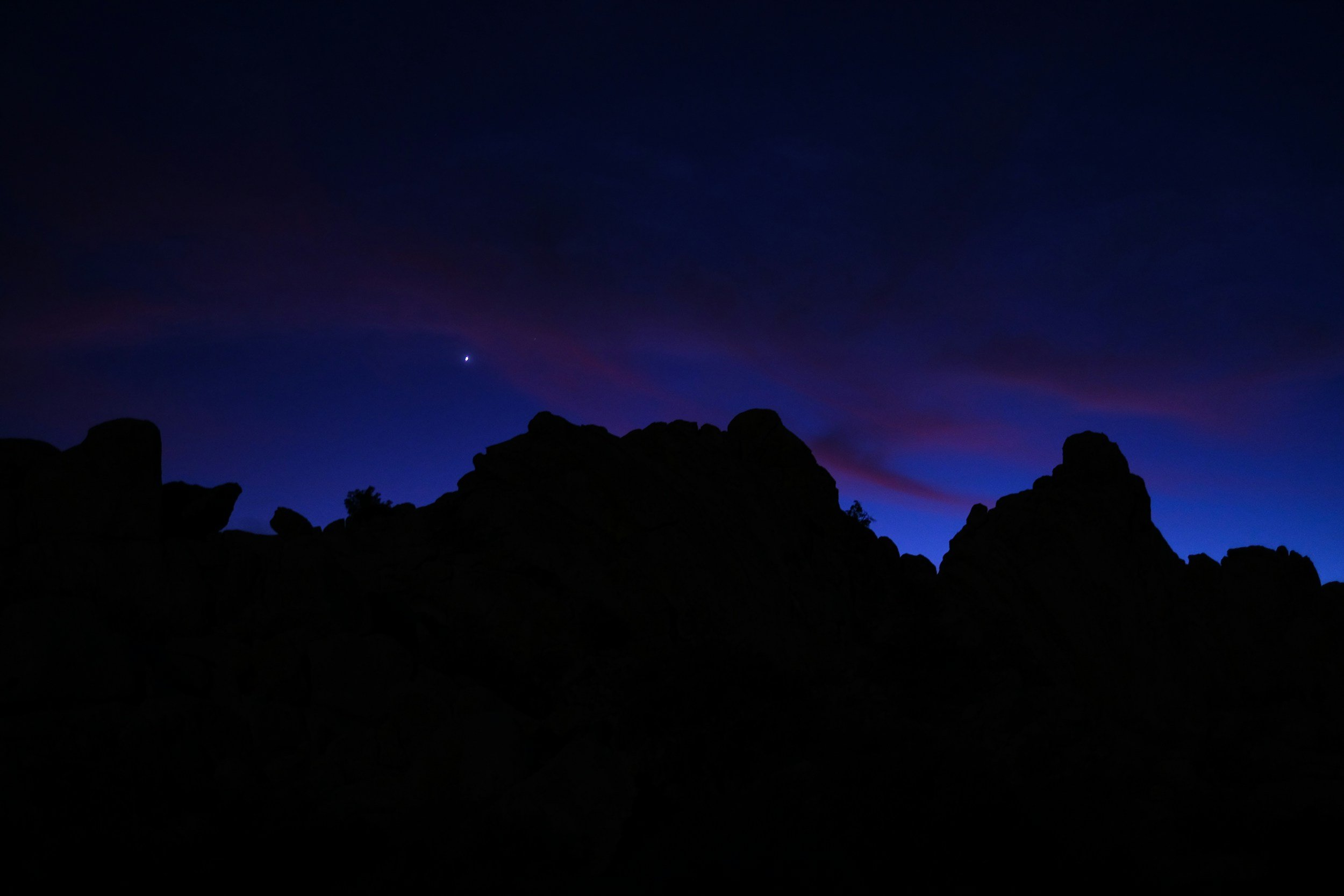 Nighttime landscape with dark silhouette of rocky mountains and a deep blue and purple sky.