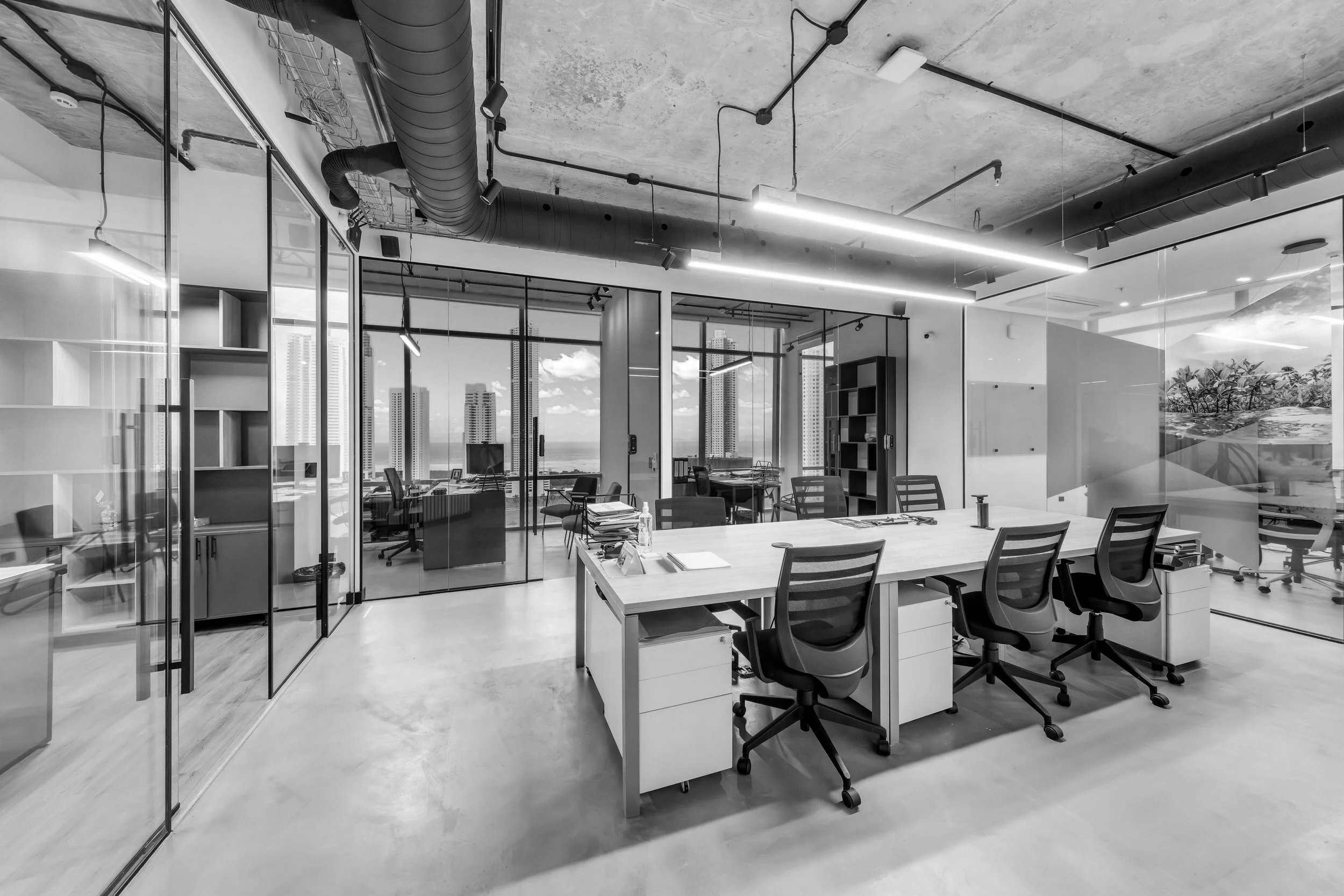 Modern office space with glass partitions, work desks and chairs, large windows showing a city skyline, exposed ceiling with ductwork, and minimalist lighting.