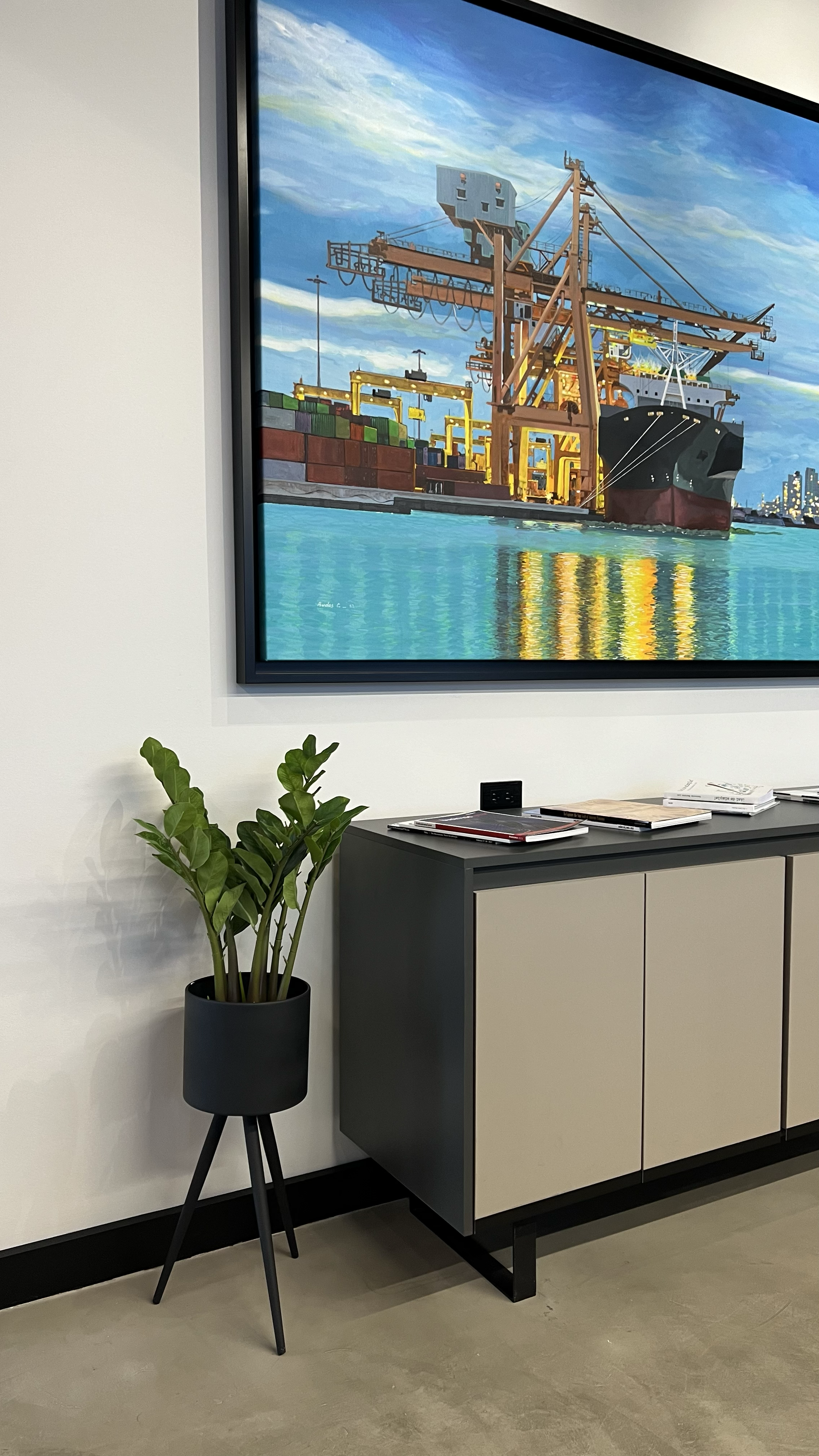 A large framed painting of an industrial harbor scene with a ship and container cranes hangs on a white wall. Below the painting, there is a modern sideboard with papers on top. To the left of the sideboard, there is a potted plant with large green l