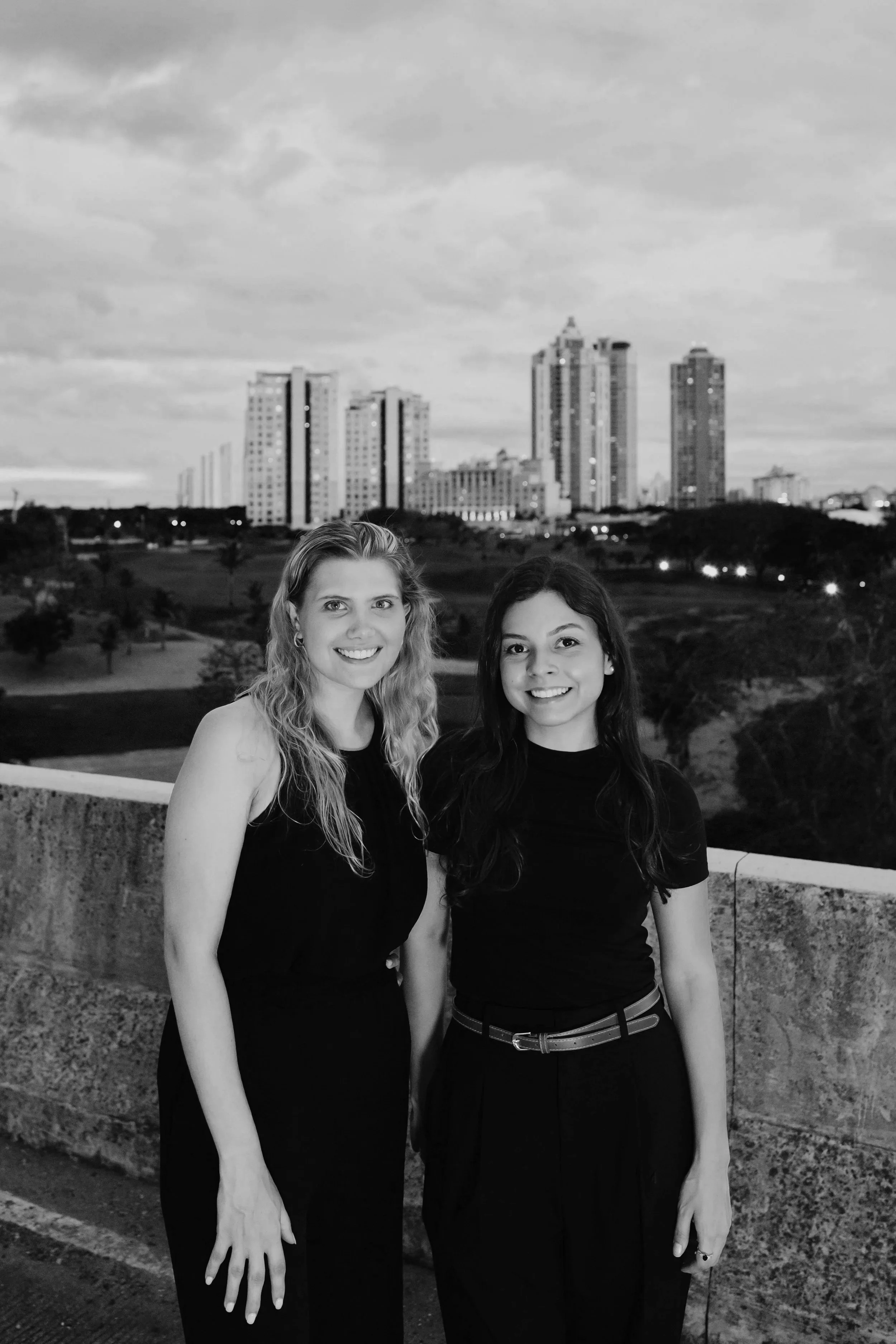 Two women standing outdoors at night with a city skyline featuring tall buildings and cloudy sky in the background.