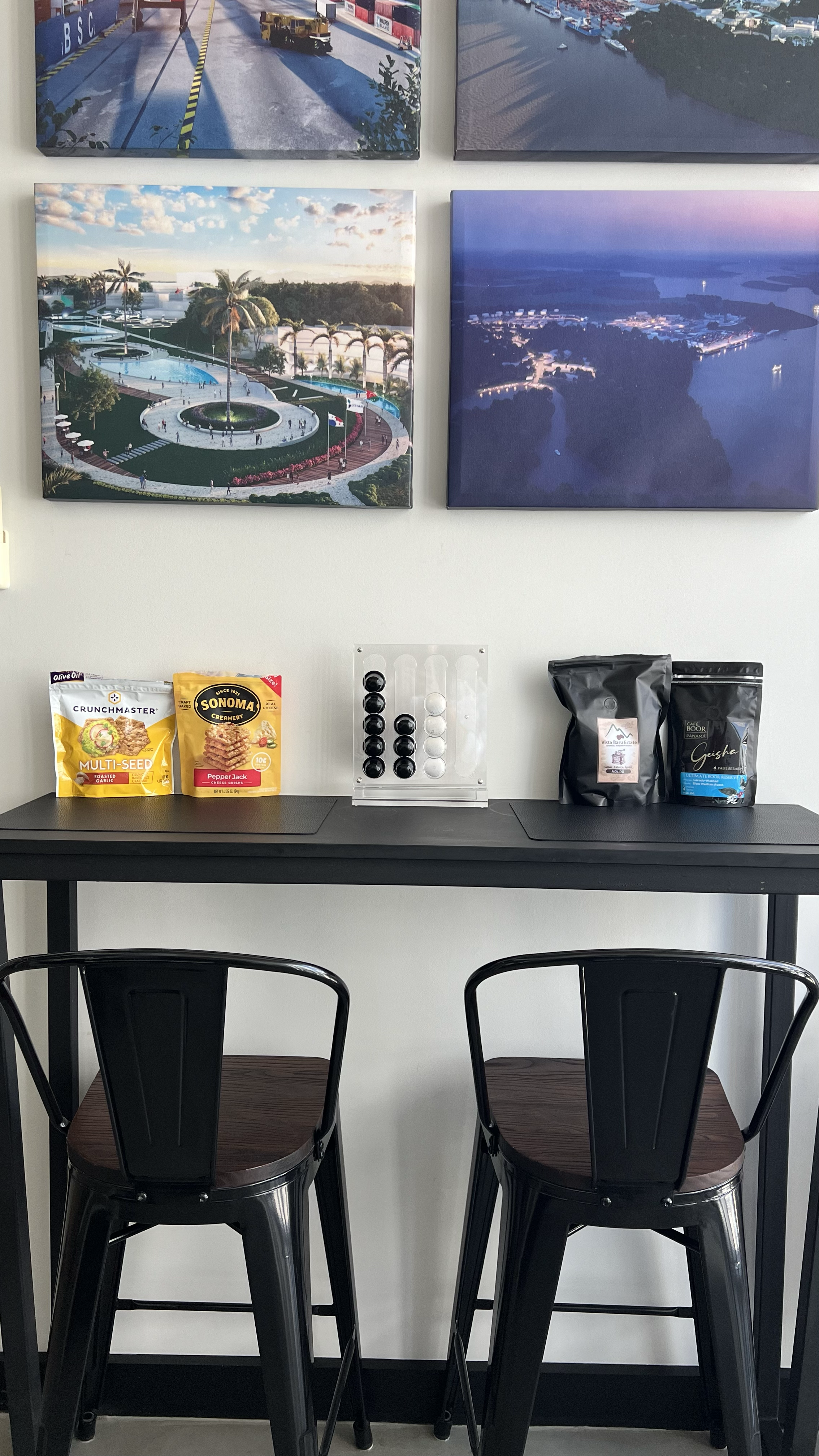 A black table with two black chairs, a white wall with four framed landscape photographs, and snacks including croutons and cheese crackers on the table.